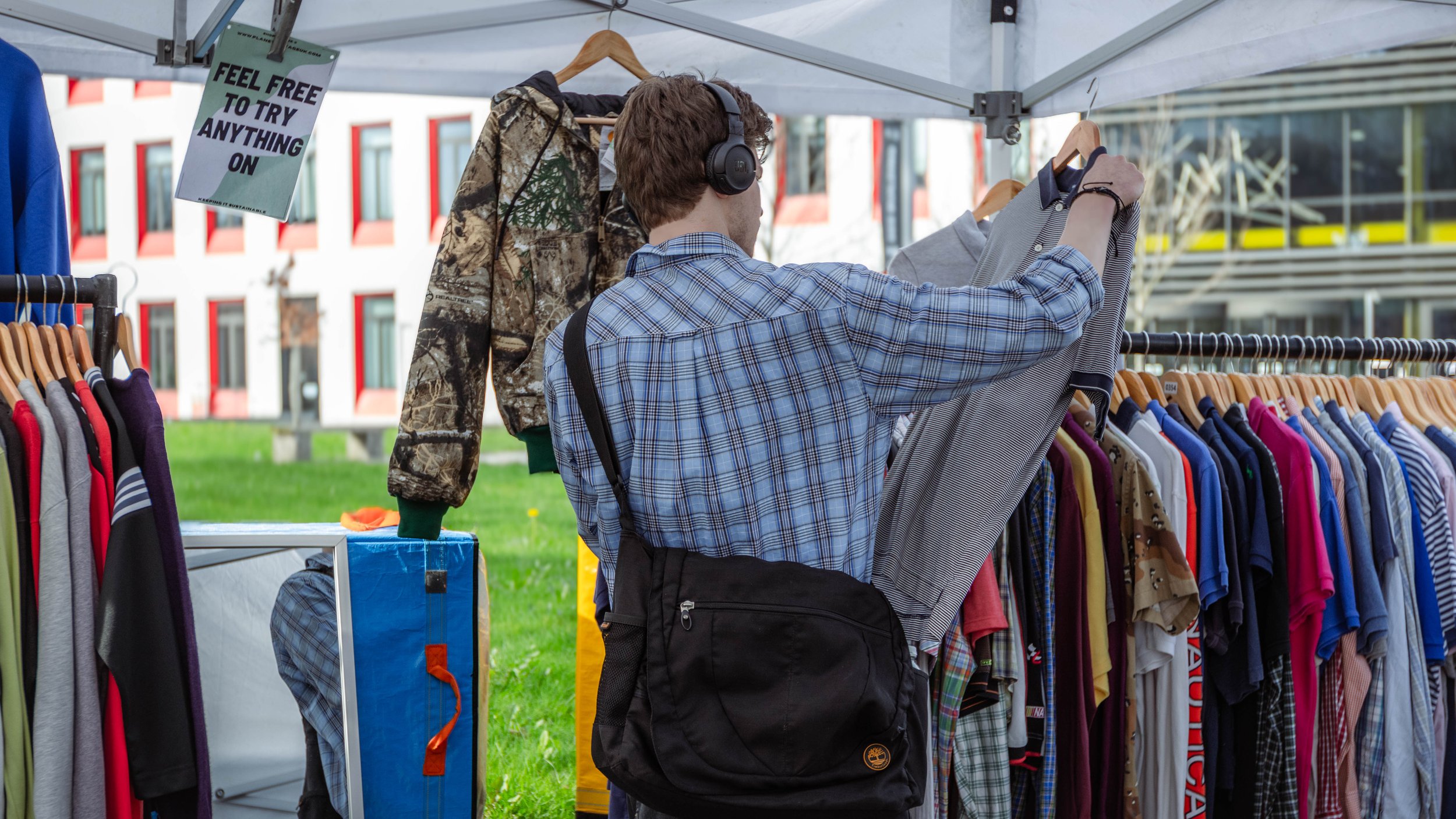 A young man shopping at an outdoor clothing market, browsing a rack of colorful shirts. He is wearing a plaid shirt, black backpack, and headphones.