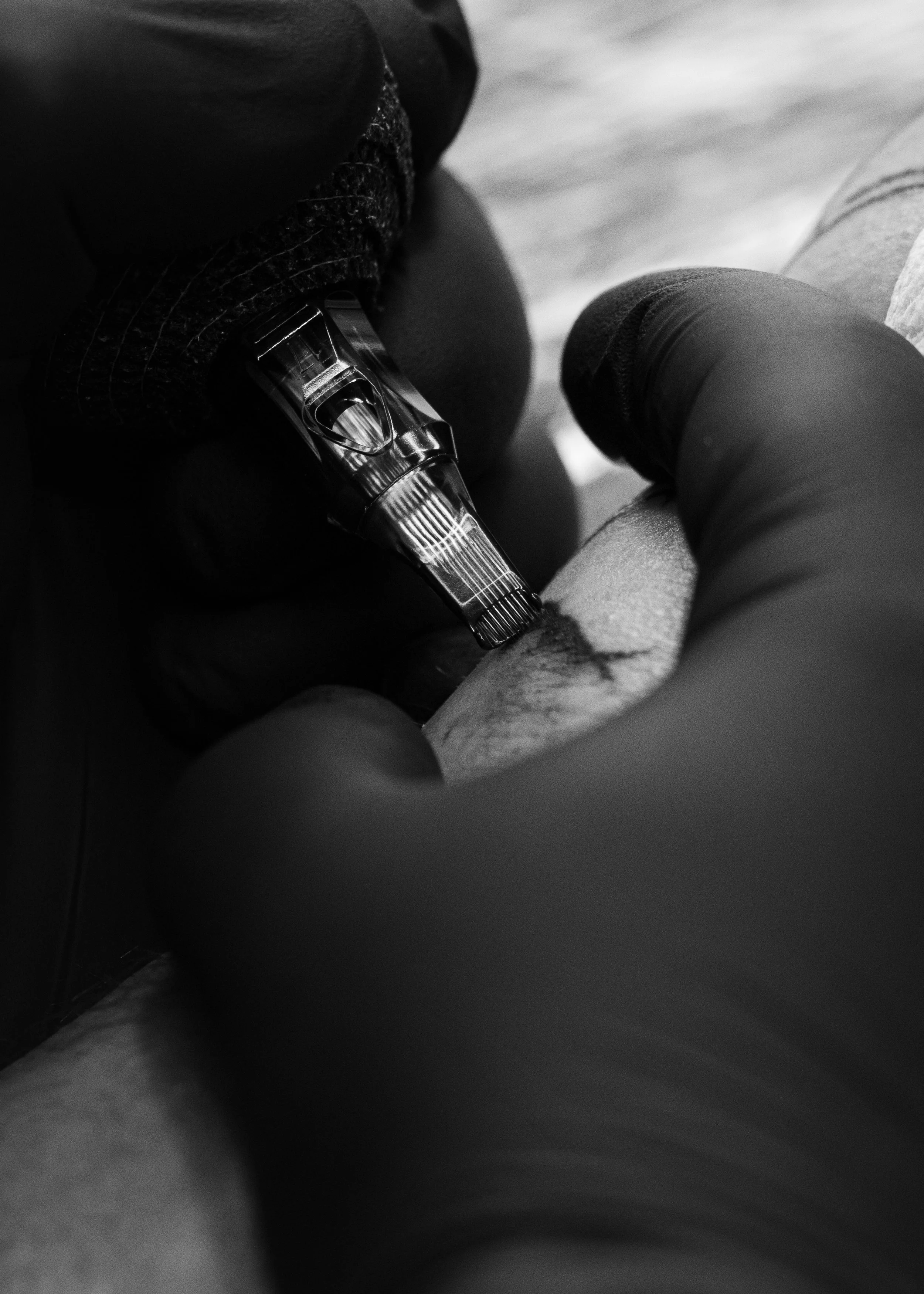 Black and white close-up image of a person's arm with a tattoo, wearing black gloves, holding a tattoo machine, tattooing skin.