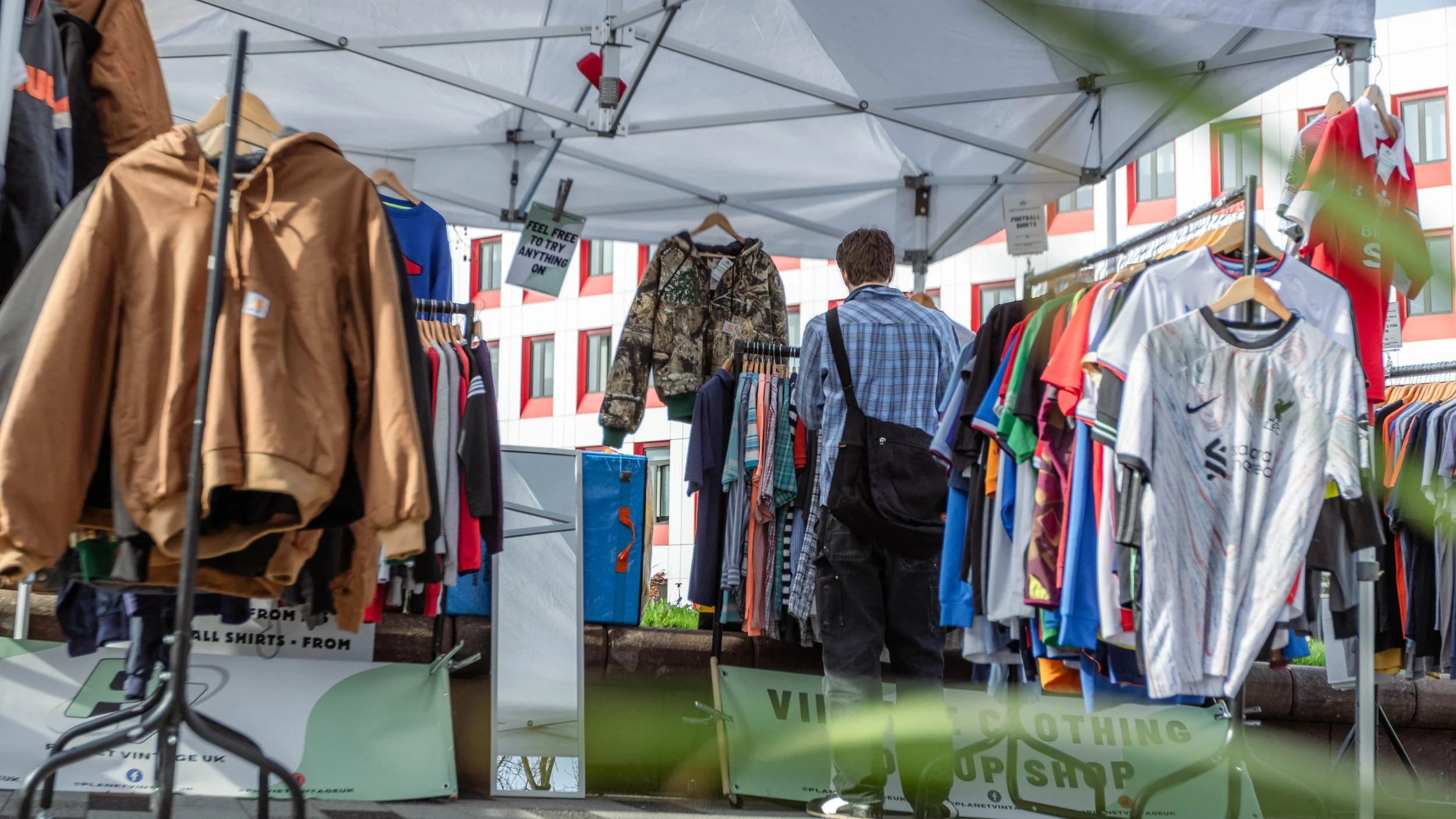 A person shopping at an outdoor clothing booth with racks of various shirts, jackets, and sportswear under a white canopy, with a modern building in the background.