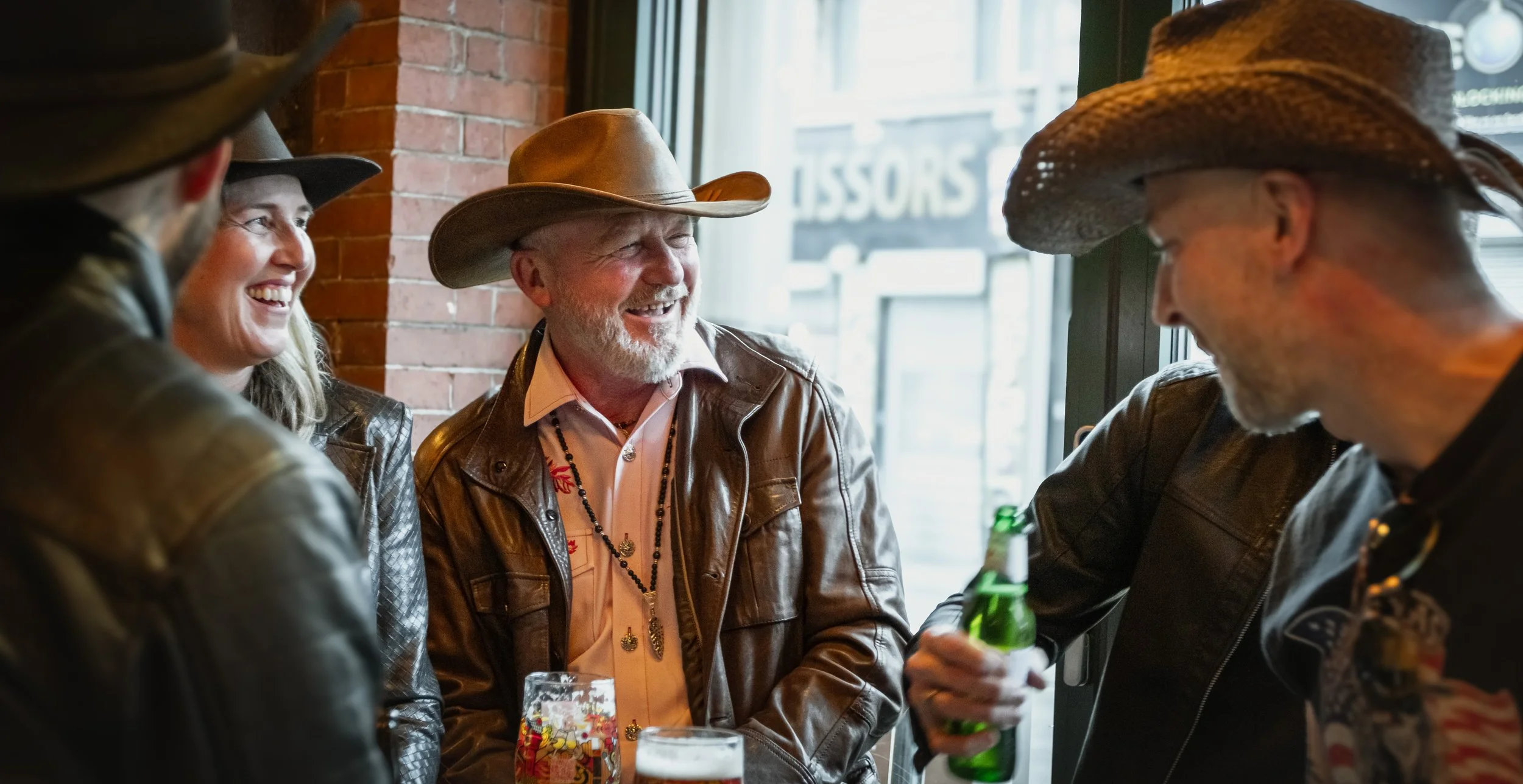 Four people with cowboy hats smiling and talking inside a bar or restaurant, with a brick wall and window in the background.
