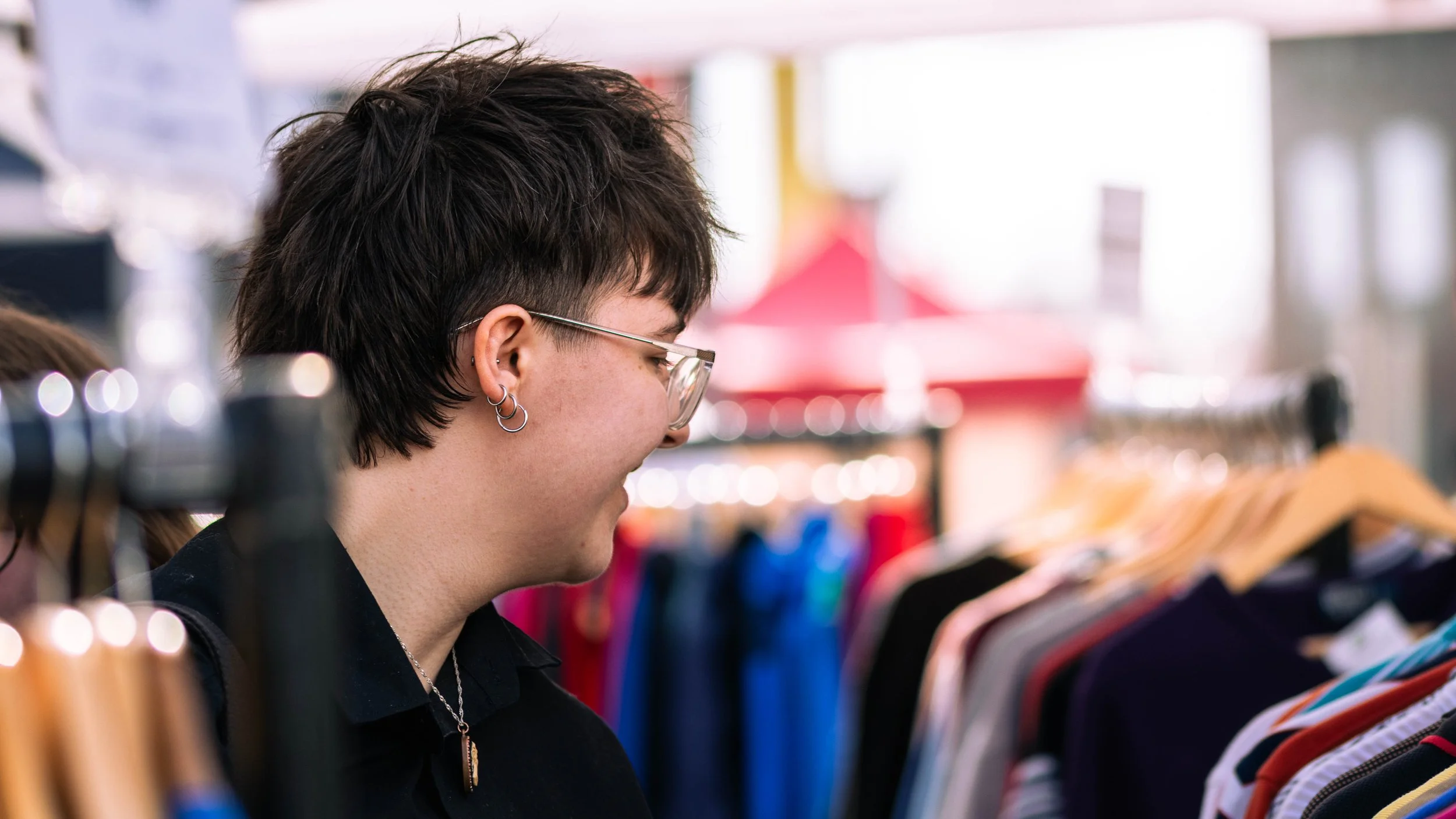Person with short dark hair, wearing glasses and earrings, shopping for clothes in a store.