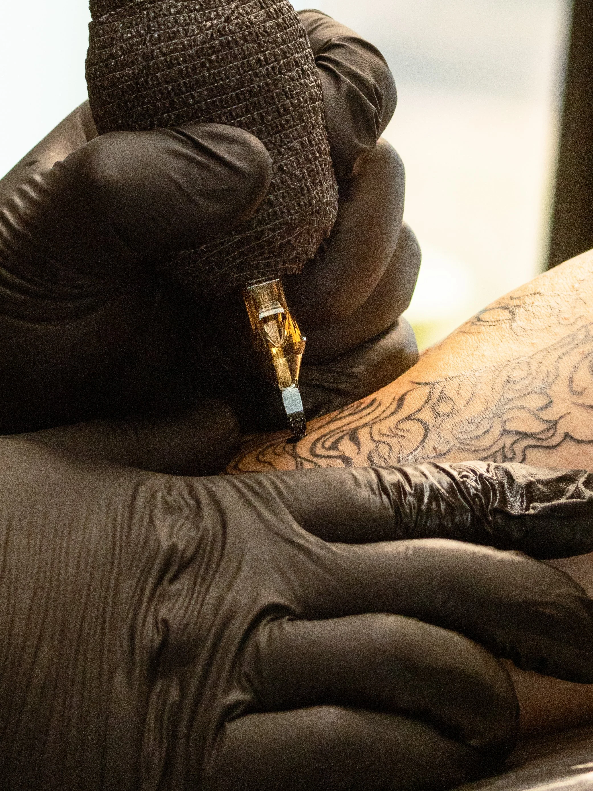 Close-up of a tattooed person's arm being tattooed by an artist using a tattoo machine. The artist wears black gloves and a brown textured shirt. The tattoo design appears to be intricate patterns or script.