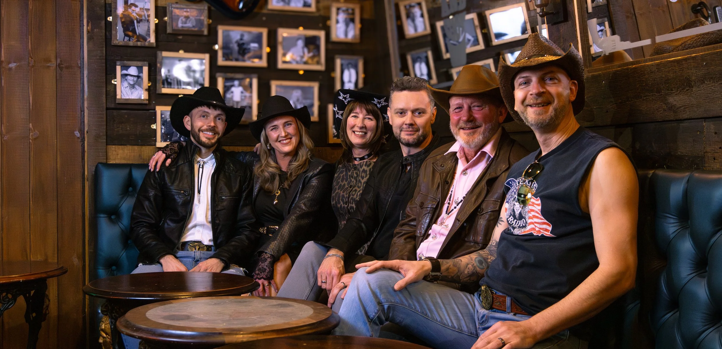 Group of six friends sitting on a booth in a bar, wearing cowboy hats and casual clothing, smiling and enjoying each other's company, with framed photos on the wooden wall behind them.