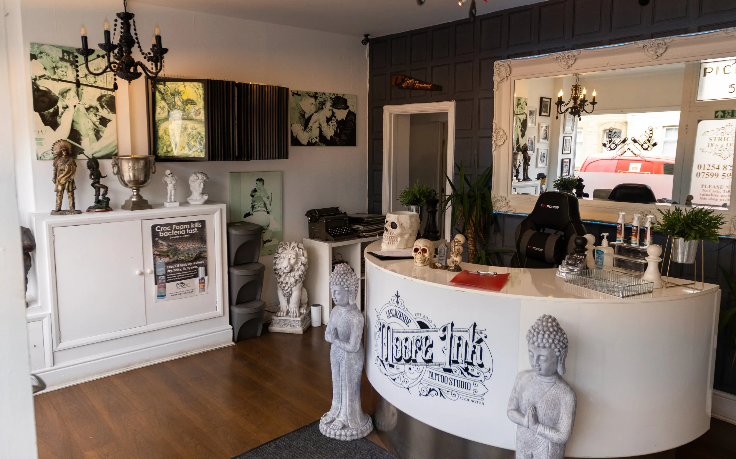 Interior of tattoo studio featuring framed photos, sculptures, and skull statues. There is a white reception desk with plants and framed art in the background.