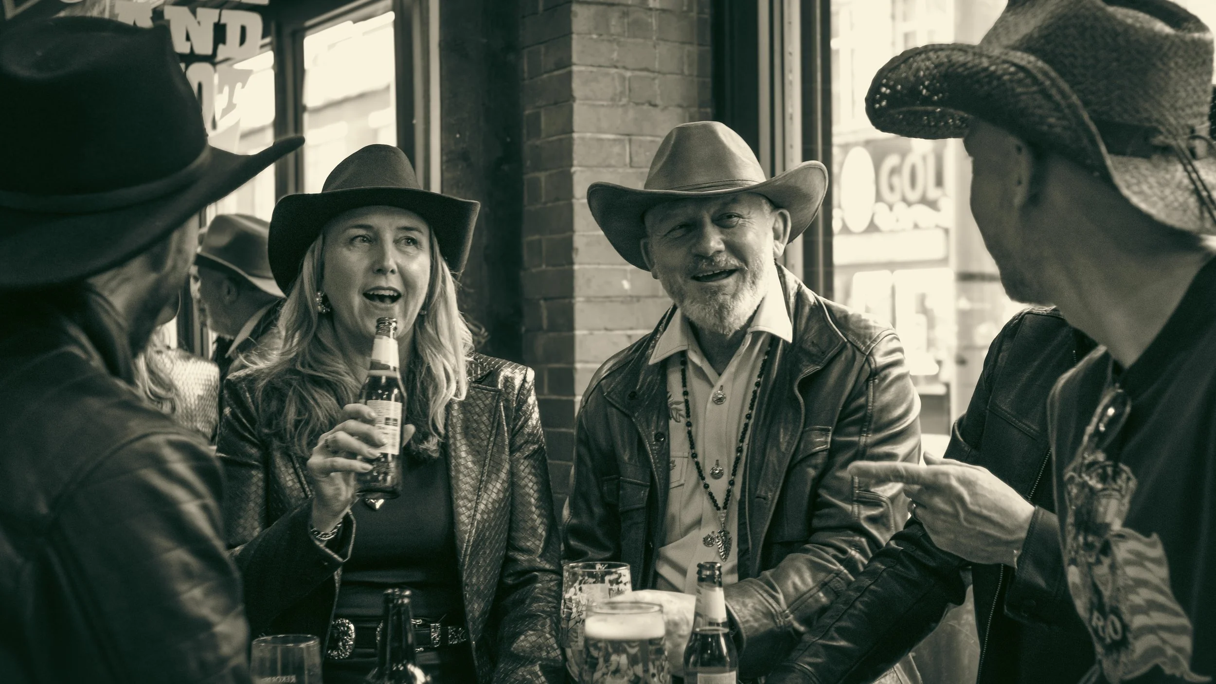 Group of people wearing cowboy hats socializing in a bar, some holding drinks, with a brick wall and windows in the background.