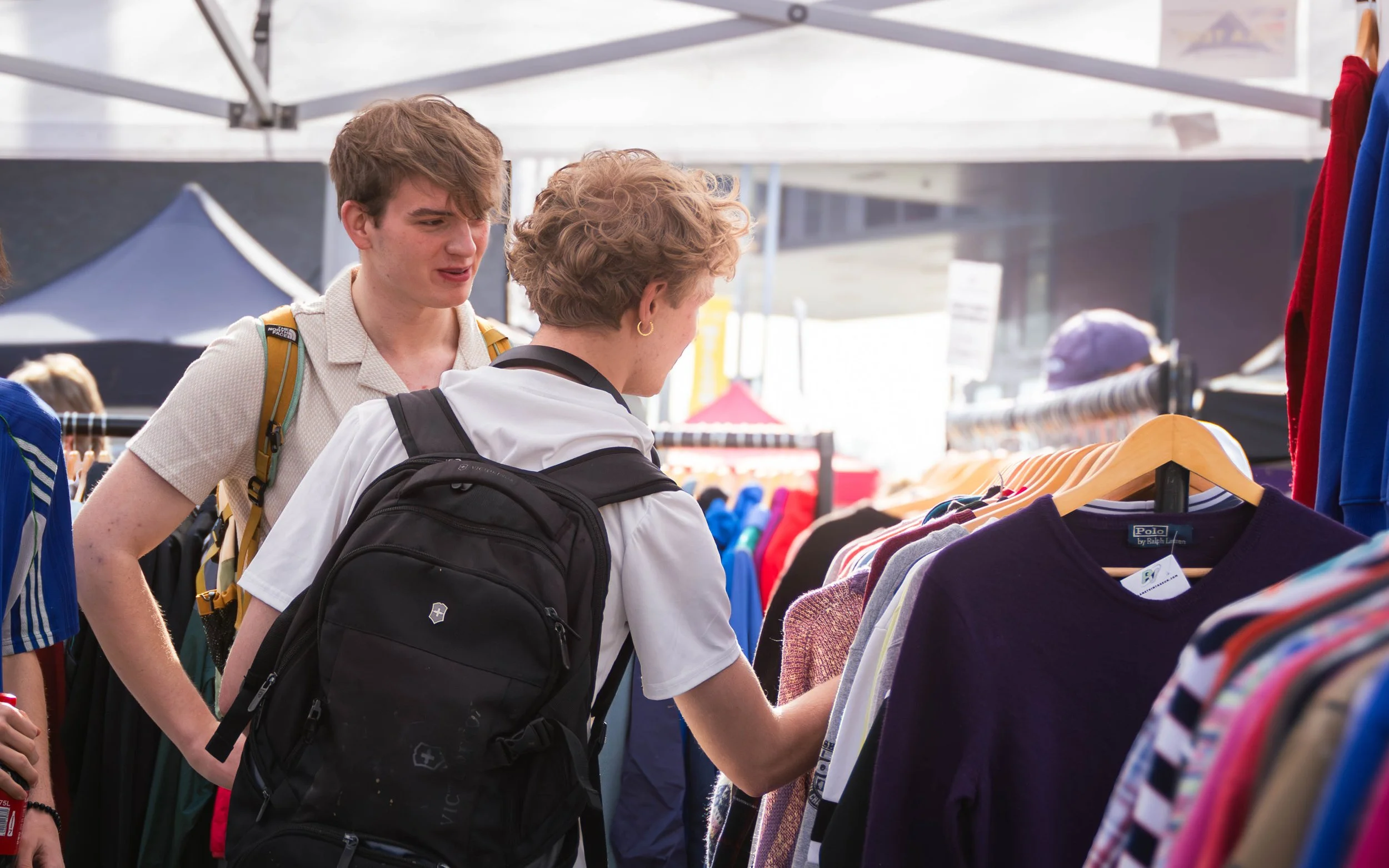 Two young boys with backpacks shopping for clothes at an outdoor market stall.