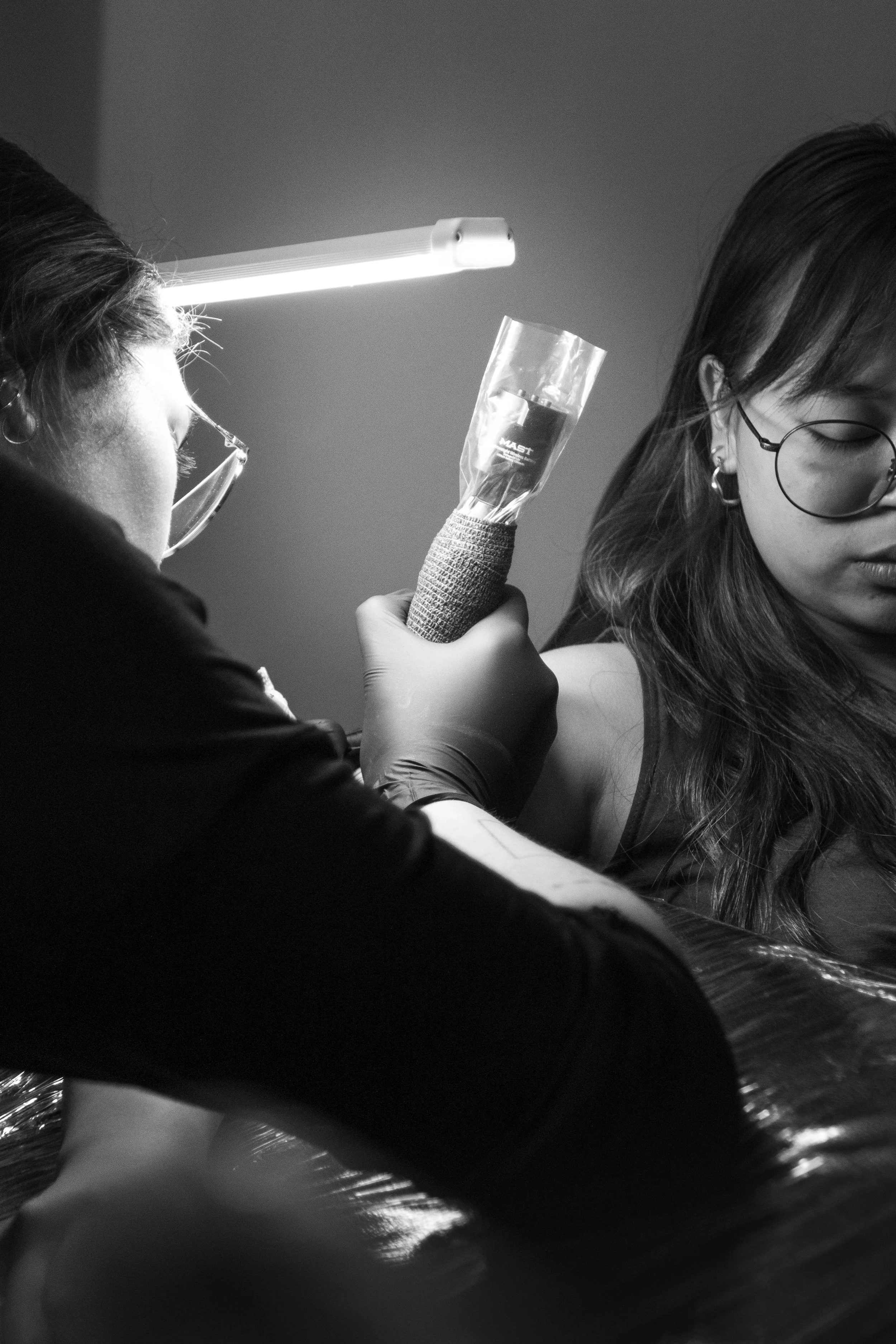 A person with long hair and glasses receives a tattoo on their shoulder from a tattoo artist. The artist is using a tattoo machine and is wearing gloves. There is a bright light overhead.