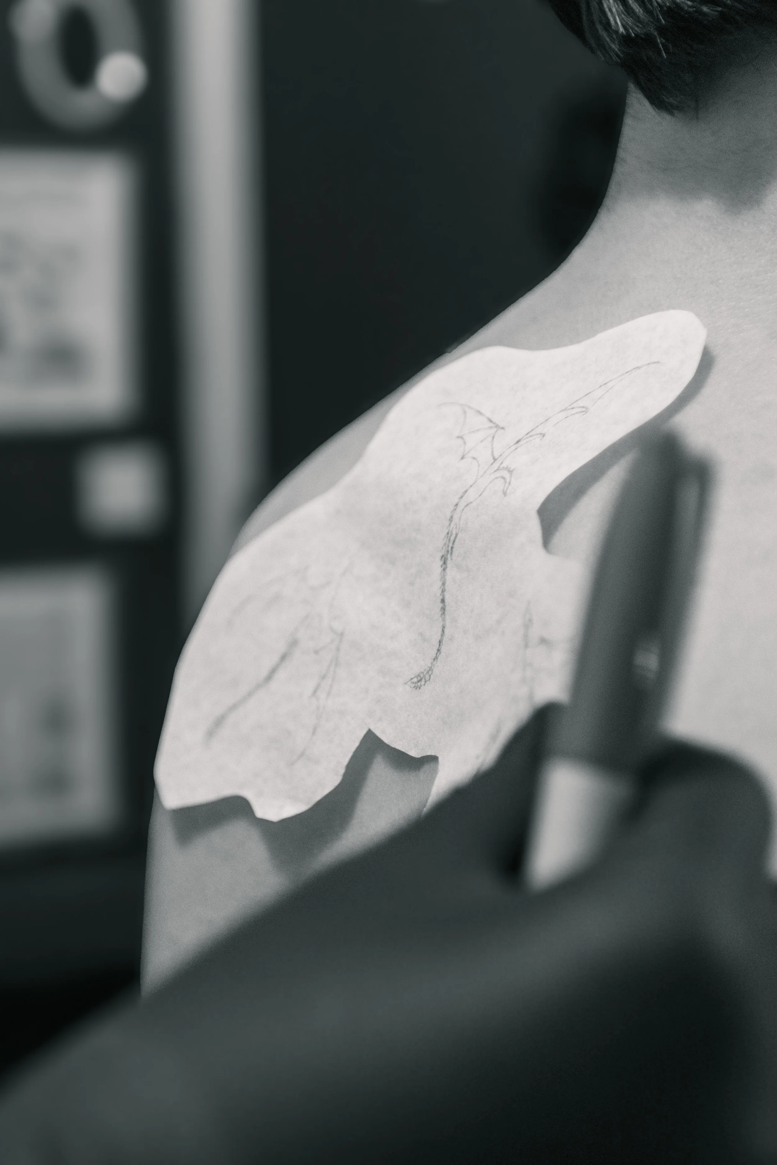 Black and white close-up photo of a hand-drawn dragon on a piece of paper attached to a surface, with a blurred view of a machine or device in the background.