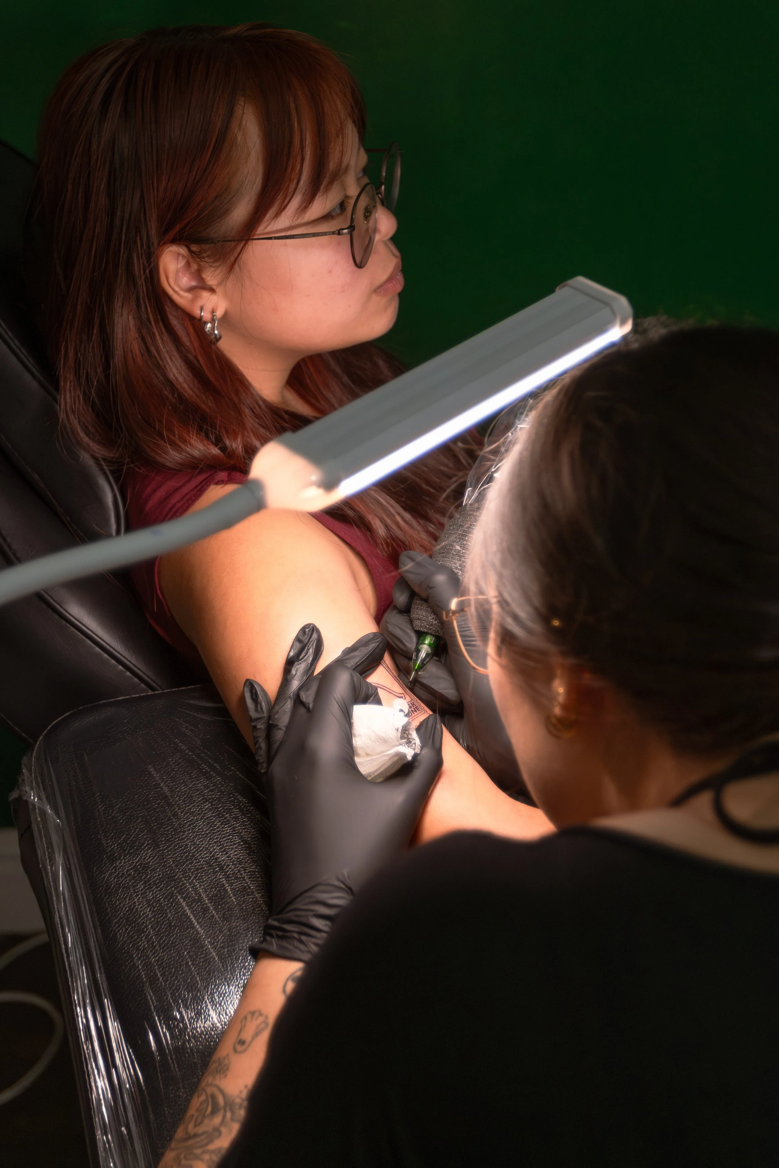 A woman with reddish hair and glasses receives a tattoo on her upper arm from a tattoo artist wearing black gloves.