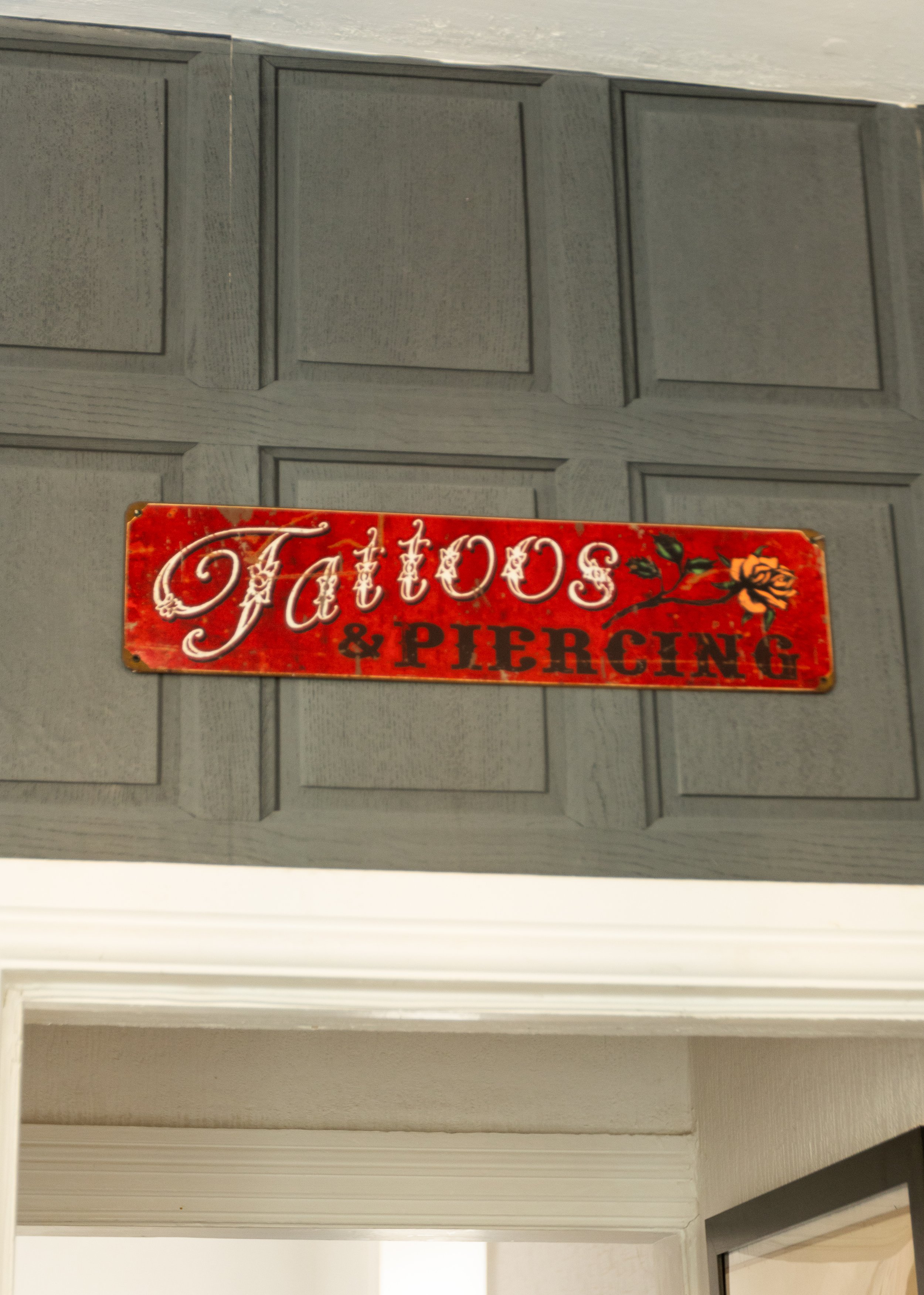 A decorative sign on a gray wooden door reads 'Tattoo & Piercing' with a rose graphic on a red background.