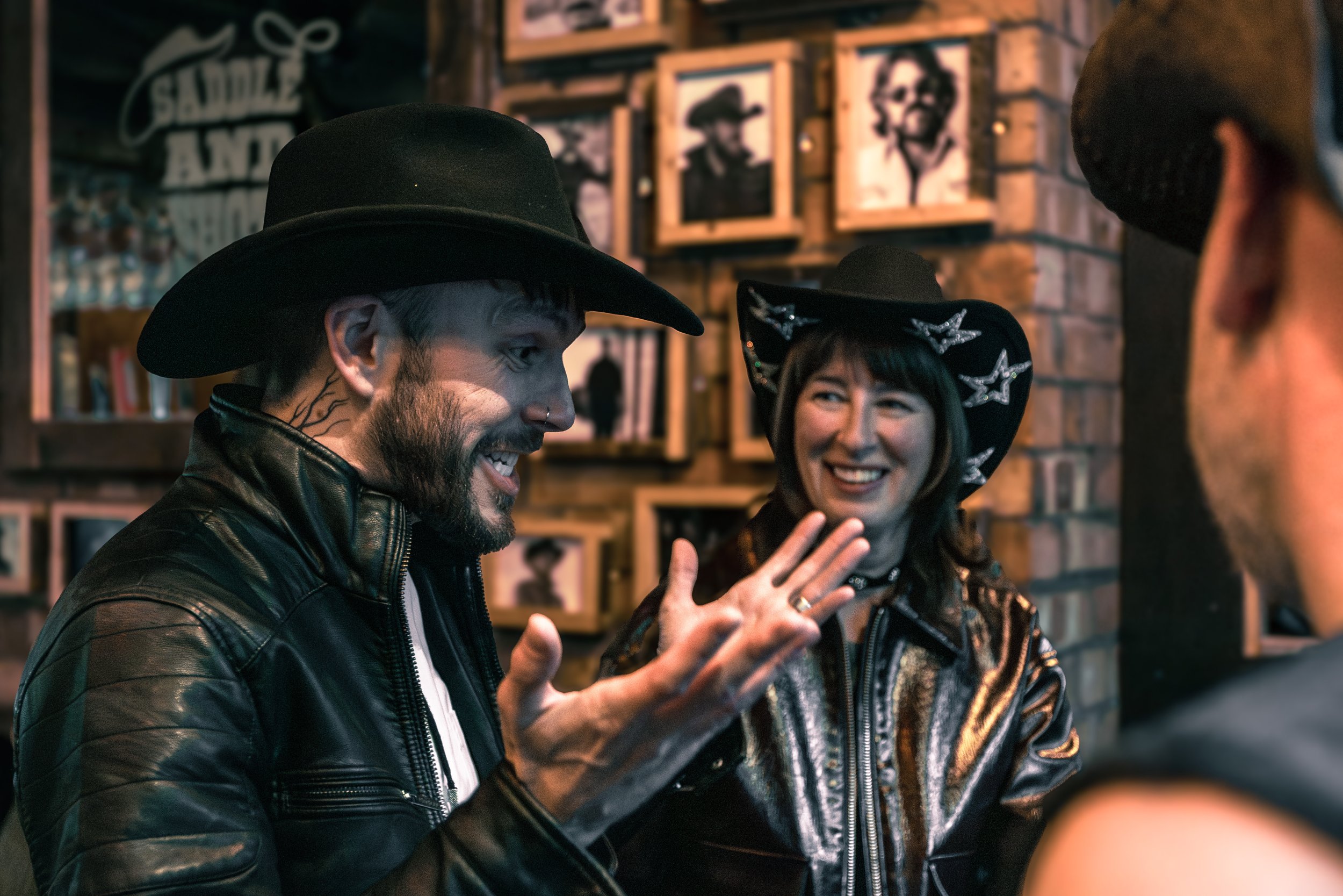 Two people in conversation at a bar or cafe, wearing leather jackets and cowboy hats, with the man smiling and gesturing with his hand, and a woman smiling and wearing a black hat with silver star decorations, in a cozy, dimly lit environment with fr