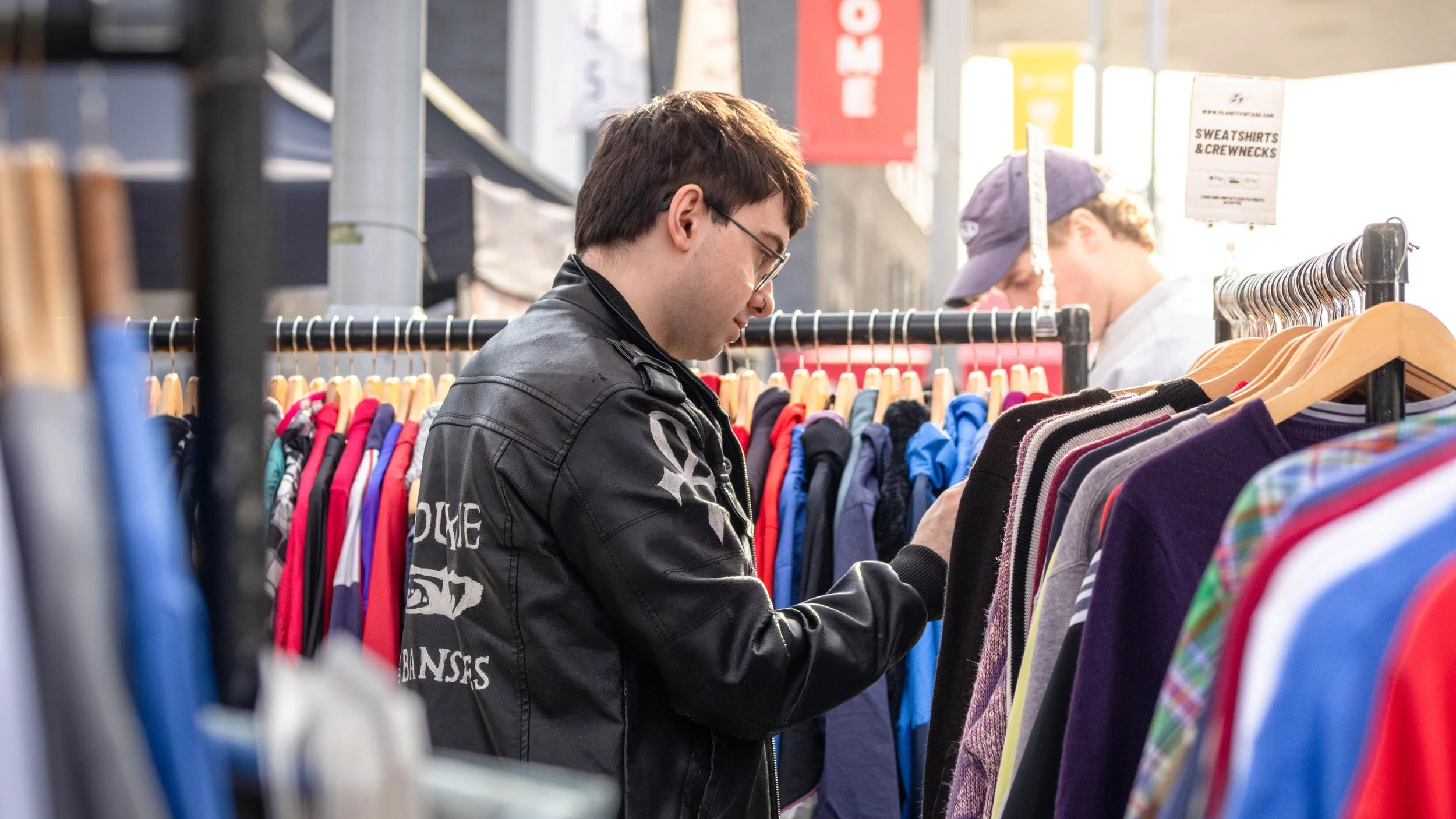 A young man with glasses and dark hair shopping at a thrift store, browsing through a rack of colorful clothes, wearing a black jacket with graphic designs.