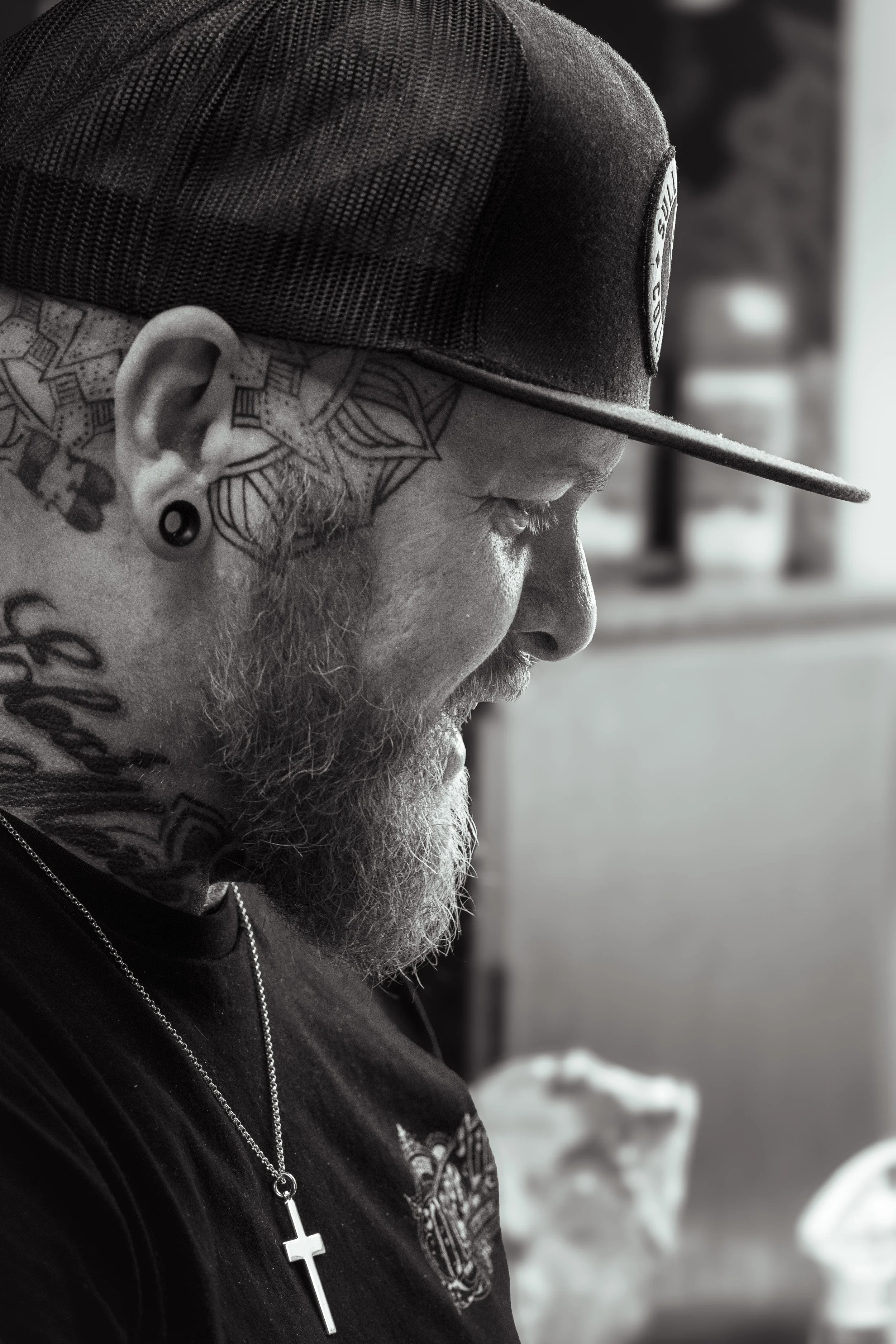 A side profile of a man with tattoos and a beard, wearing a black baseball cap and a cross necklace, standing outdoors in winter.