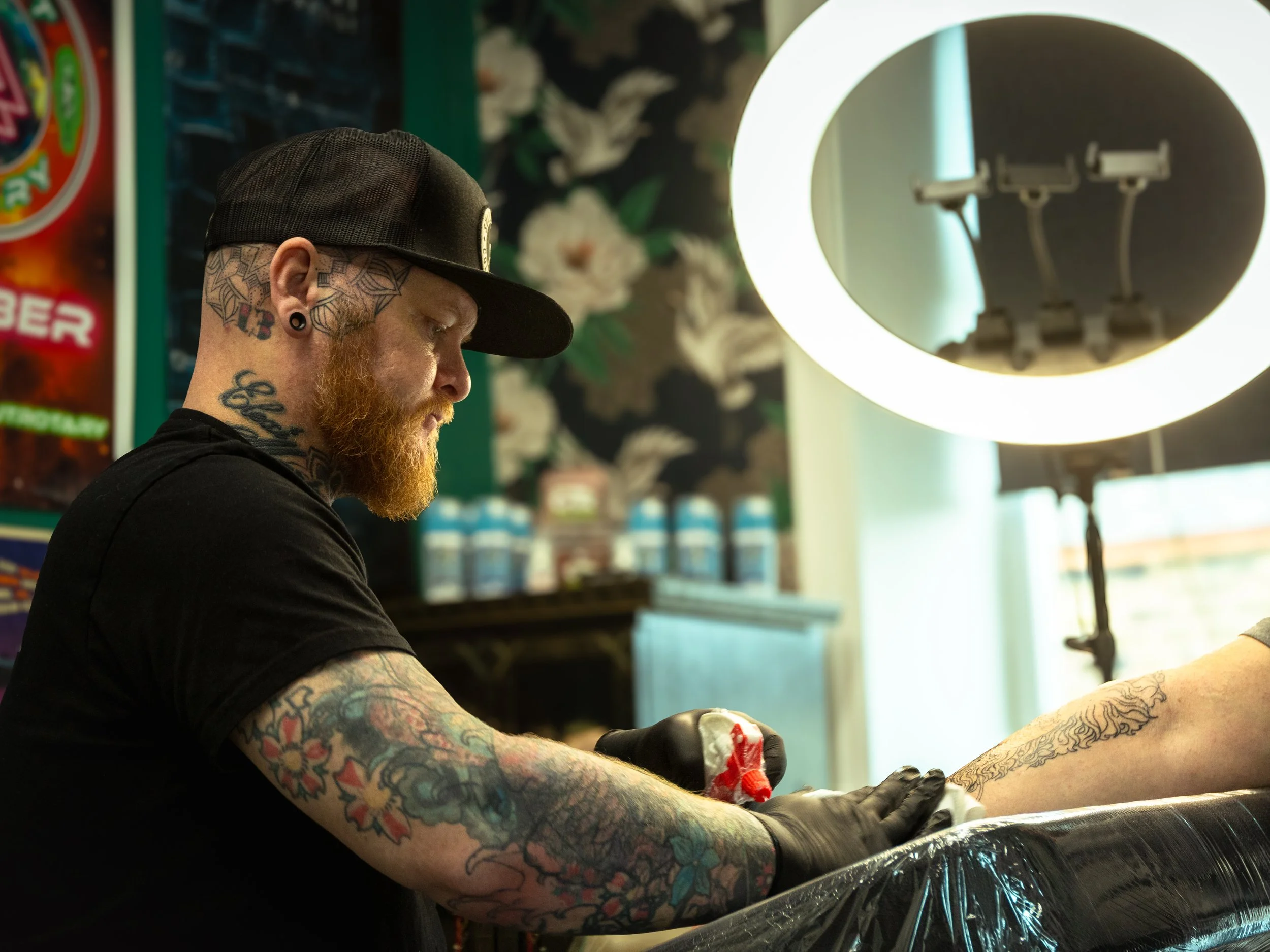 A tattooed man with a red beard, black cap, and gauge earrings tattoos a person's arm in a tattoo shop, illuminated by a bright ring light.