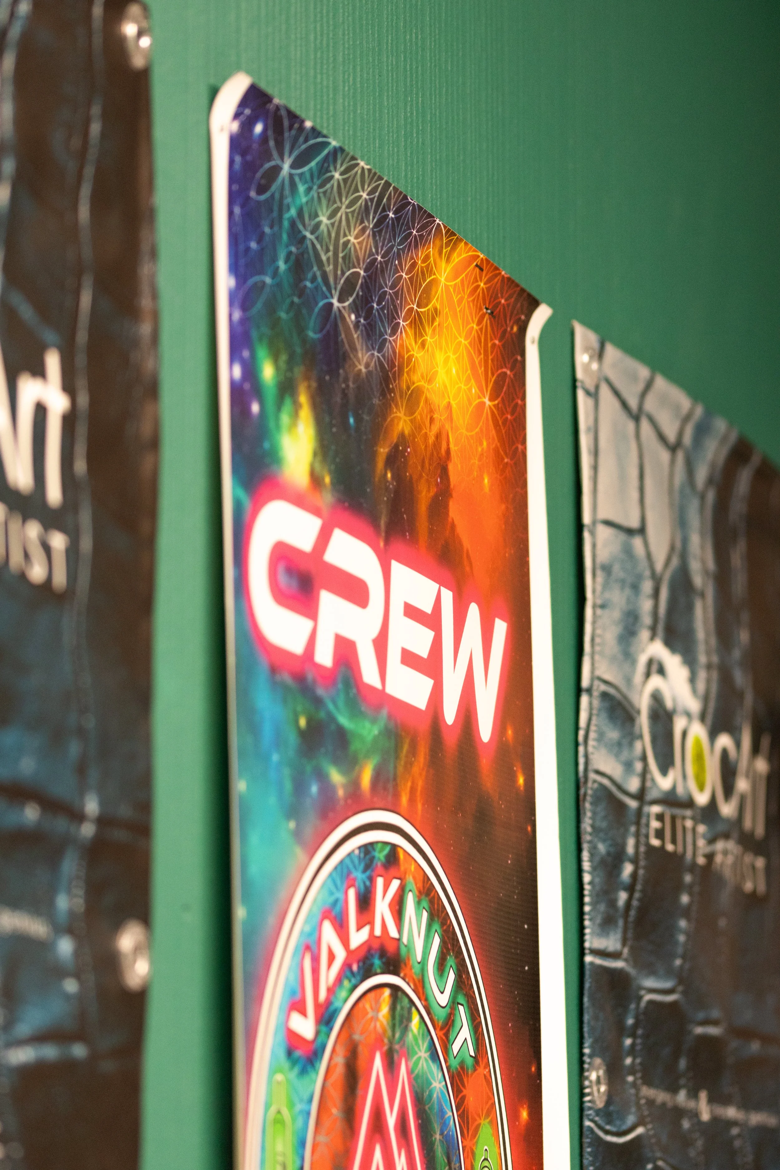Close-up of a colorful round sticker or sign with a cosmic or psychedelic design, mounted on a green wall with parts of other posters or signs visible on either side.