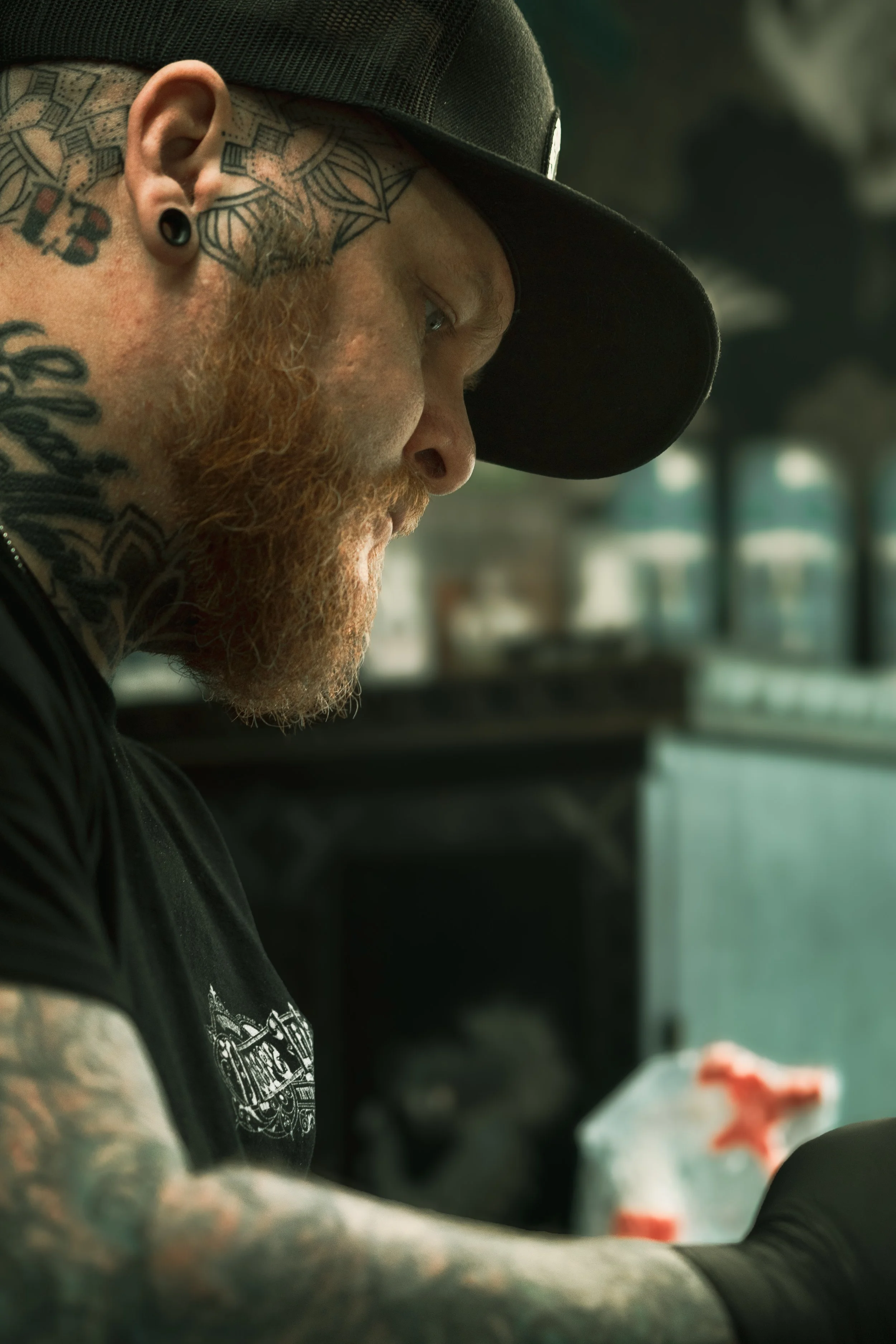 A close-up profile of a man with a red beard, tattoos on his neck and arm, wearing a black cap and earring, looking down at something in his hands.