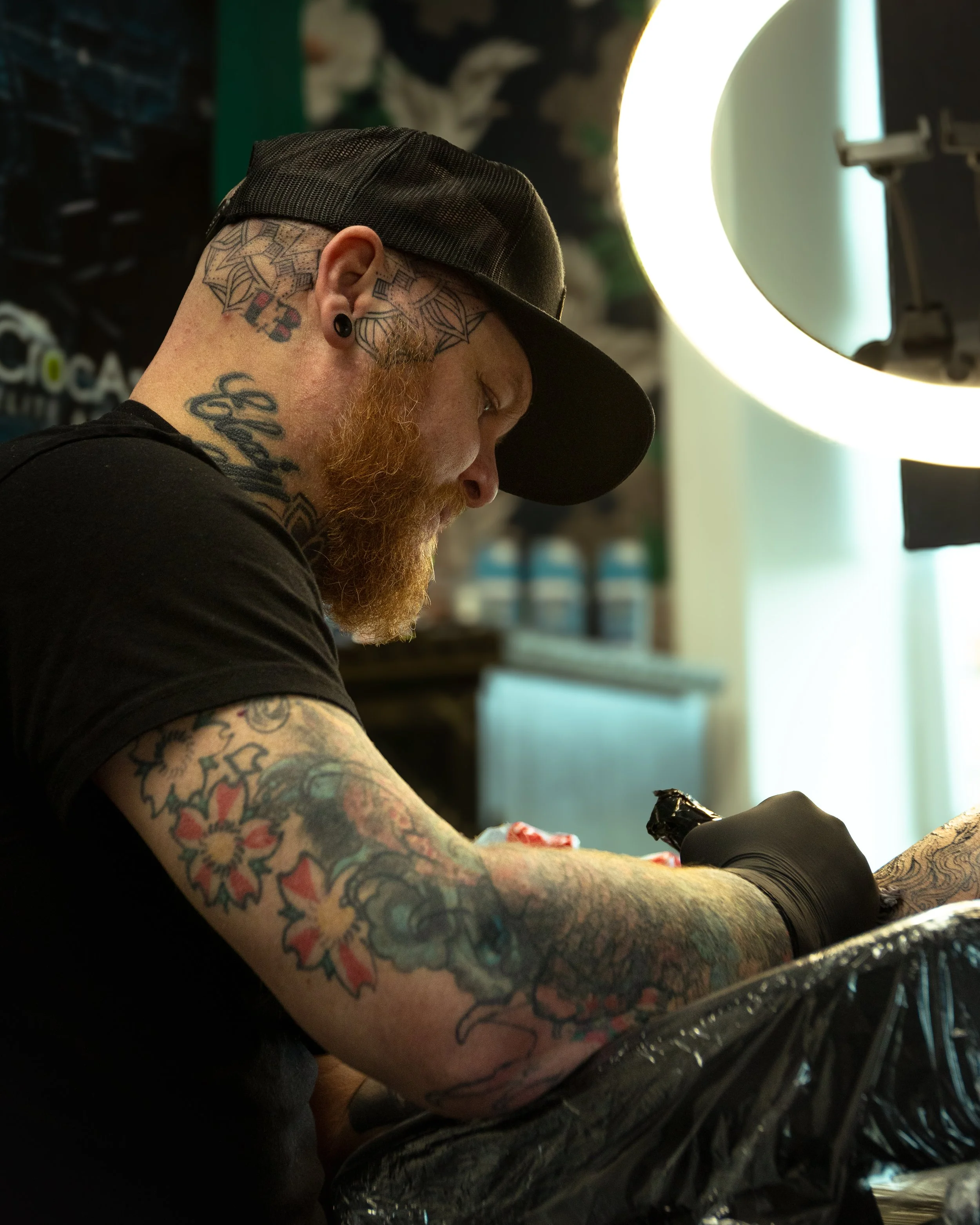 A person with tattoos on their neck and arm, wearing a black cap and black gloves, is tattooing in a tattoo studio with bright ring light and tattoo equipment visible.