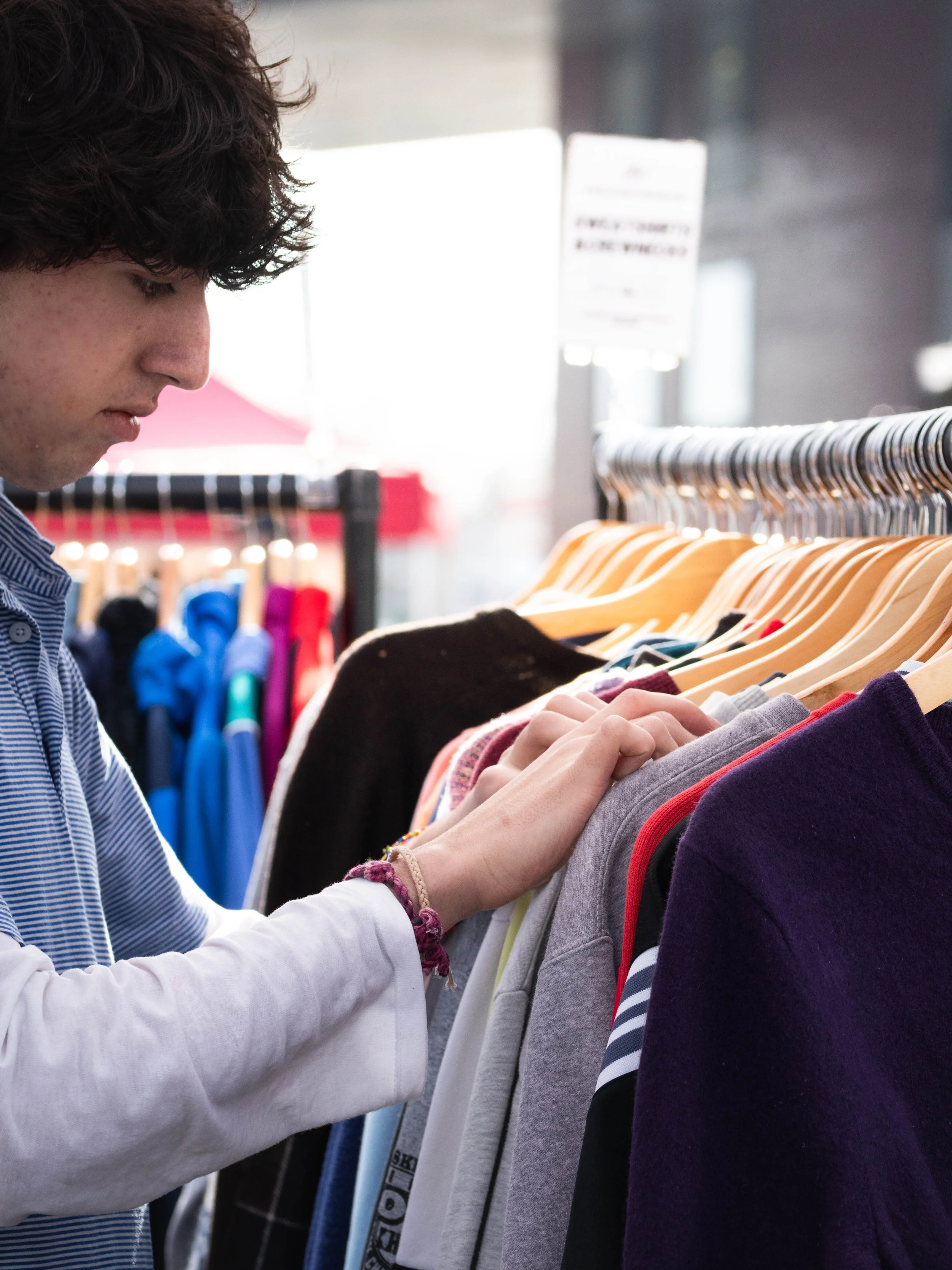 A person with dark, curly hair wearing a striped shirt and white sleeves looking at and touching clothes on a rack in a store.