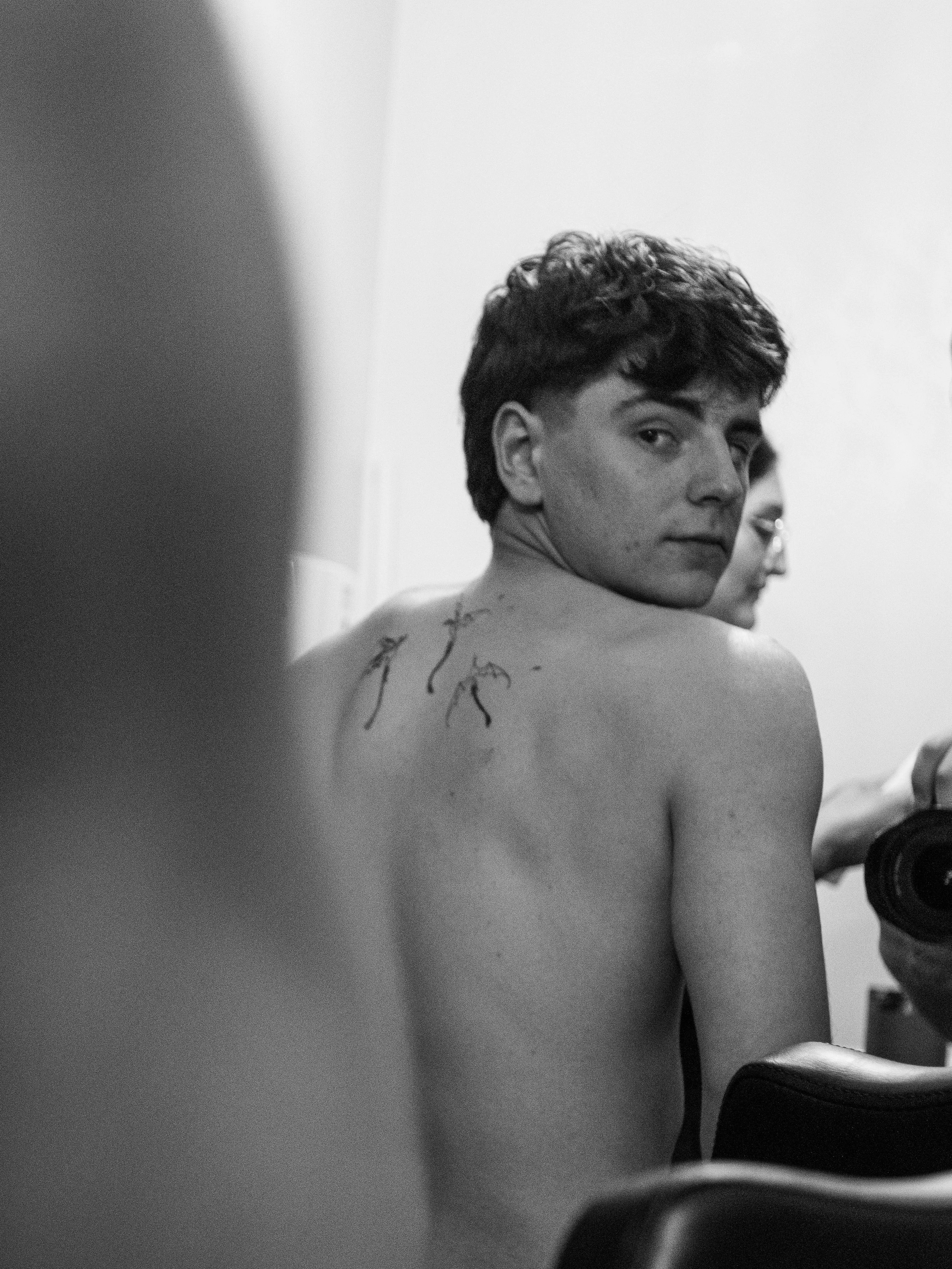Black and white photo of a young man with curly hair looking over his shoulder, with a partial view of another person in the background and a blurred foreground on the left.