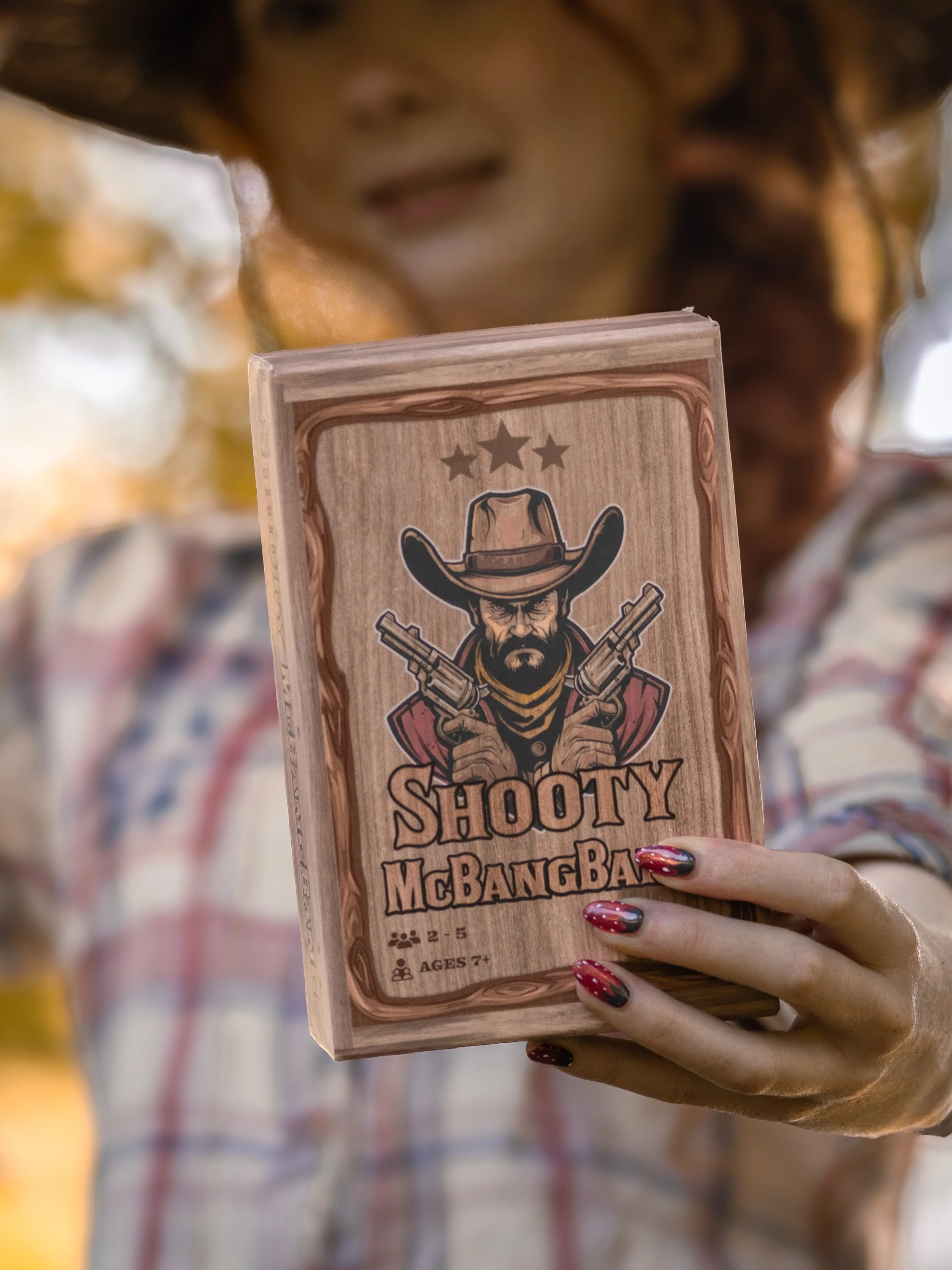 Person holding a wooden card with a cowboy graphic and text that reads 'Shooty McBangBang'.