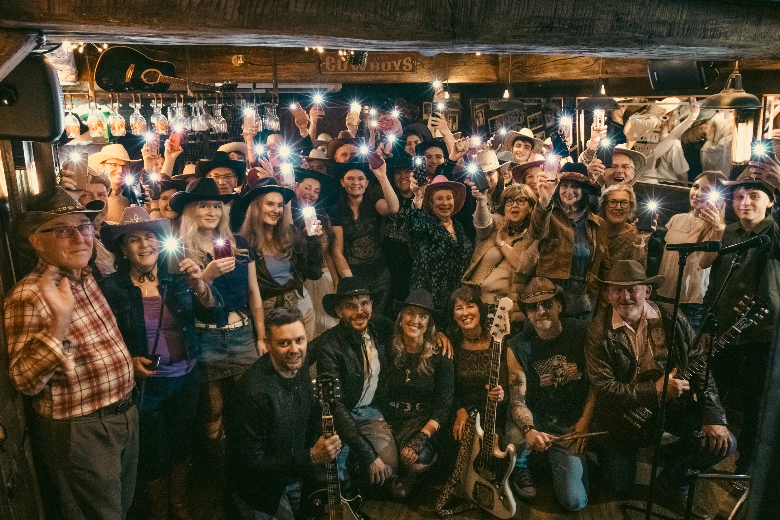 A large group of people wearing cowboy hats gathered in a dimly lit bar or restaurant, with some holding smartphones displaying flashlights, and musical instruments like guitars visible. The background has a sign that says 'LIVE COUNTRY BOYS' and han
