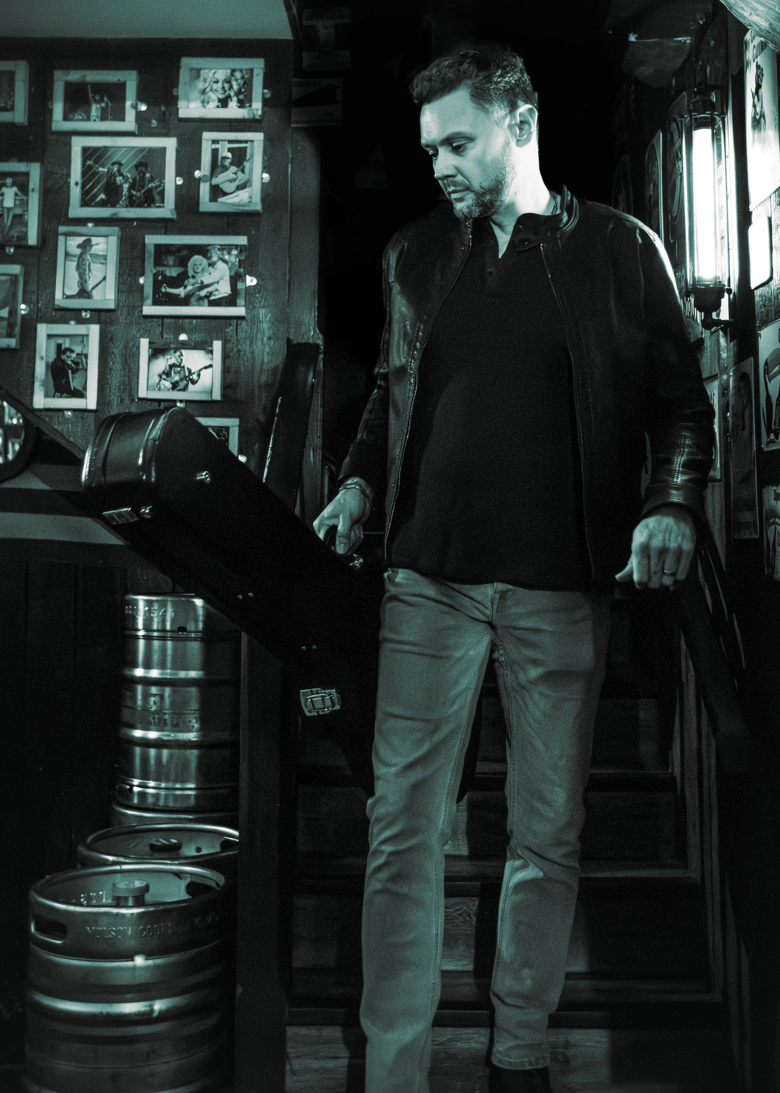 Man with short hair and beard wearing a black leather jacket and beige pants, standing indoors near a window with a wall of photos in the background, and beer kegs on the floor.