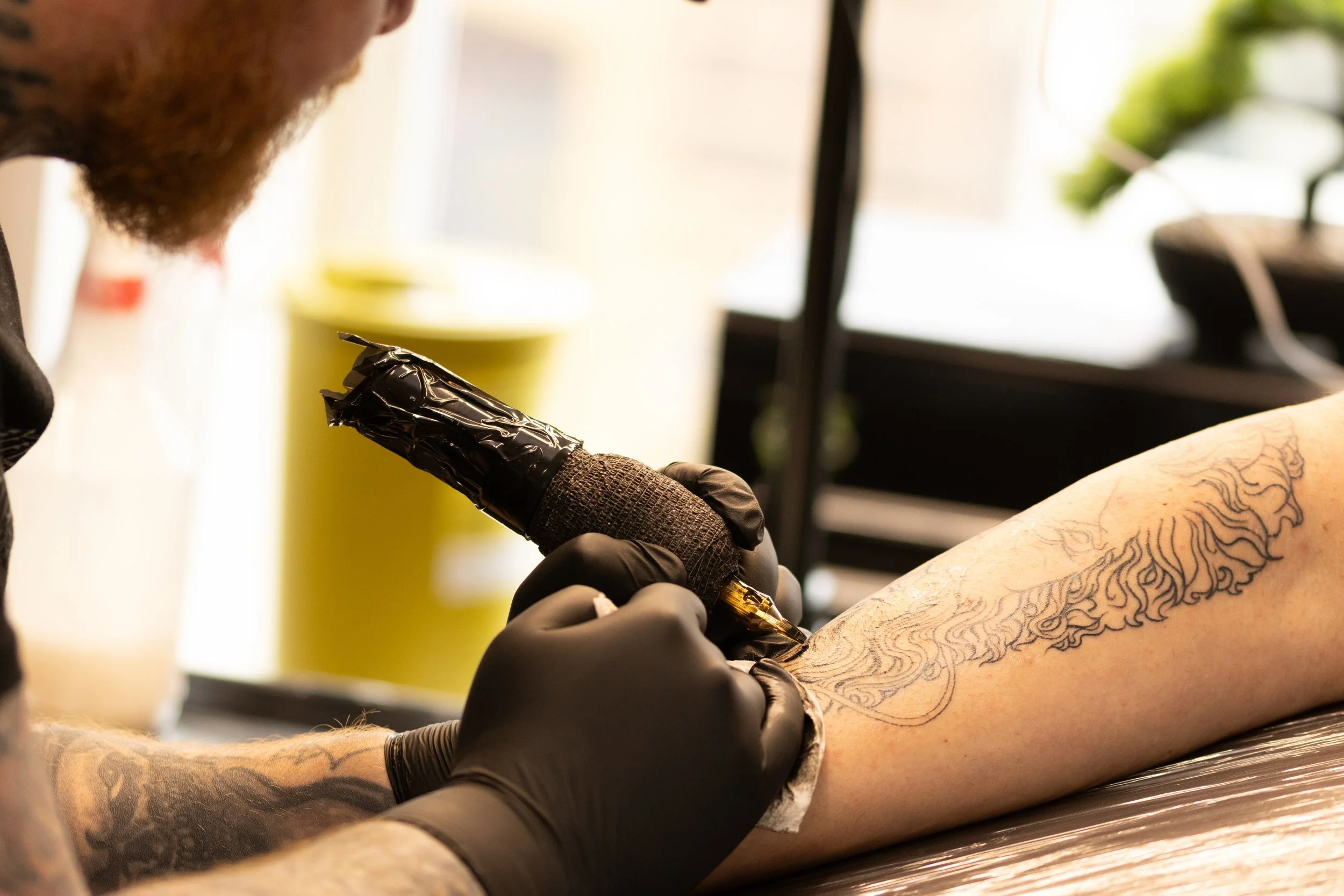 Tattoo artist tattooing a detailed design on a person's arm in a studio with various equipment and greenery visible in the background.