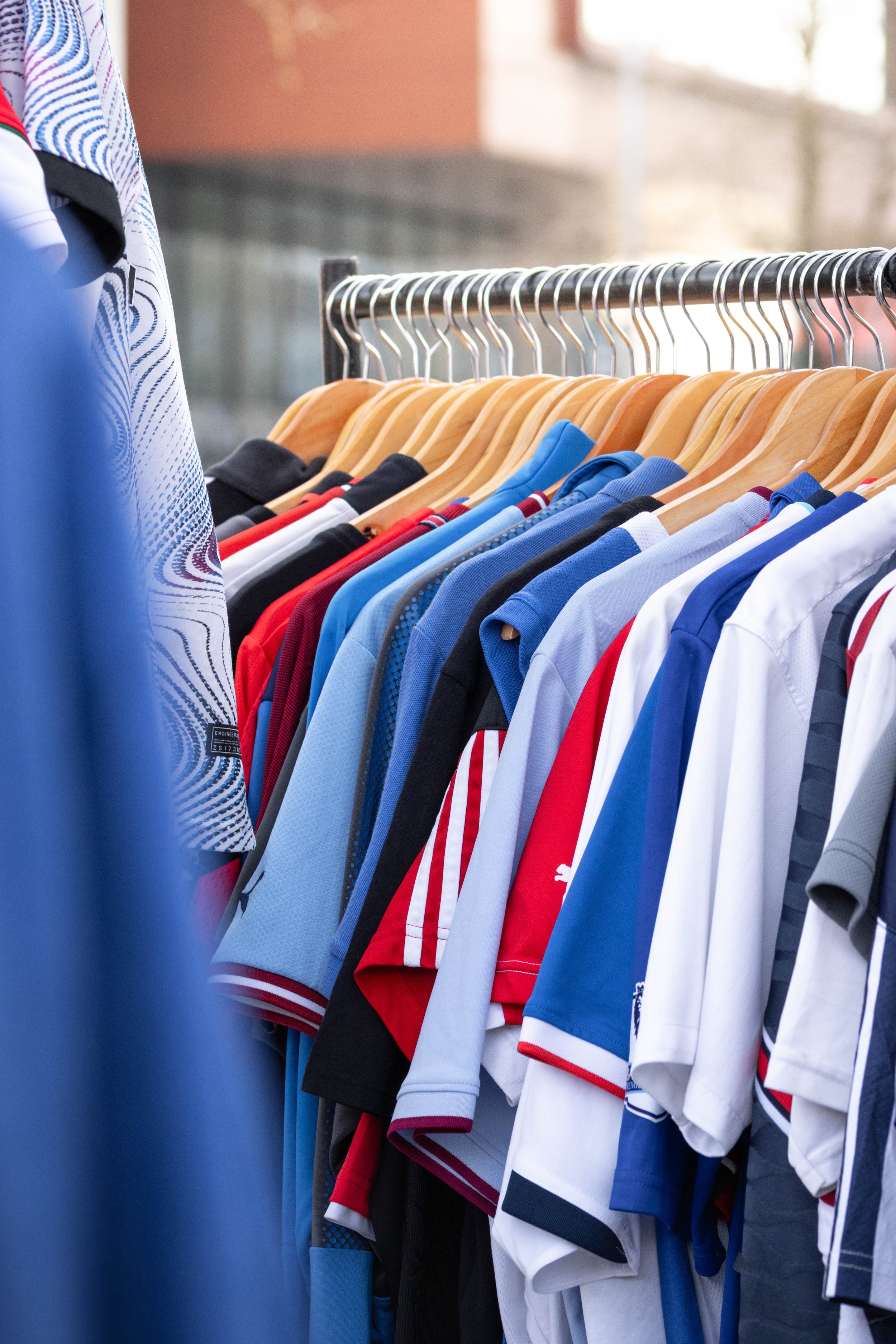 Clothing rack with sports jerseys and shirts in various colors, including blue, red, black, and white, hanging on wooden hangers.