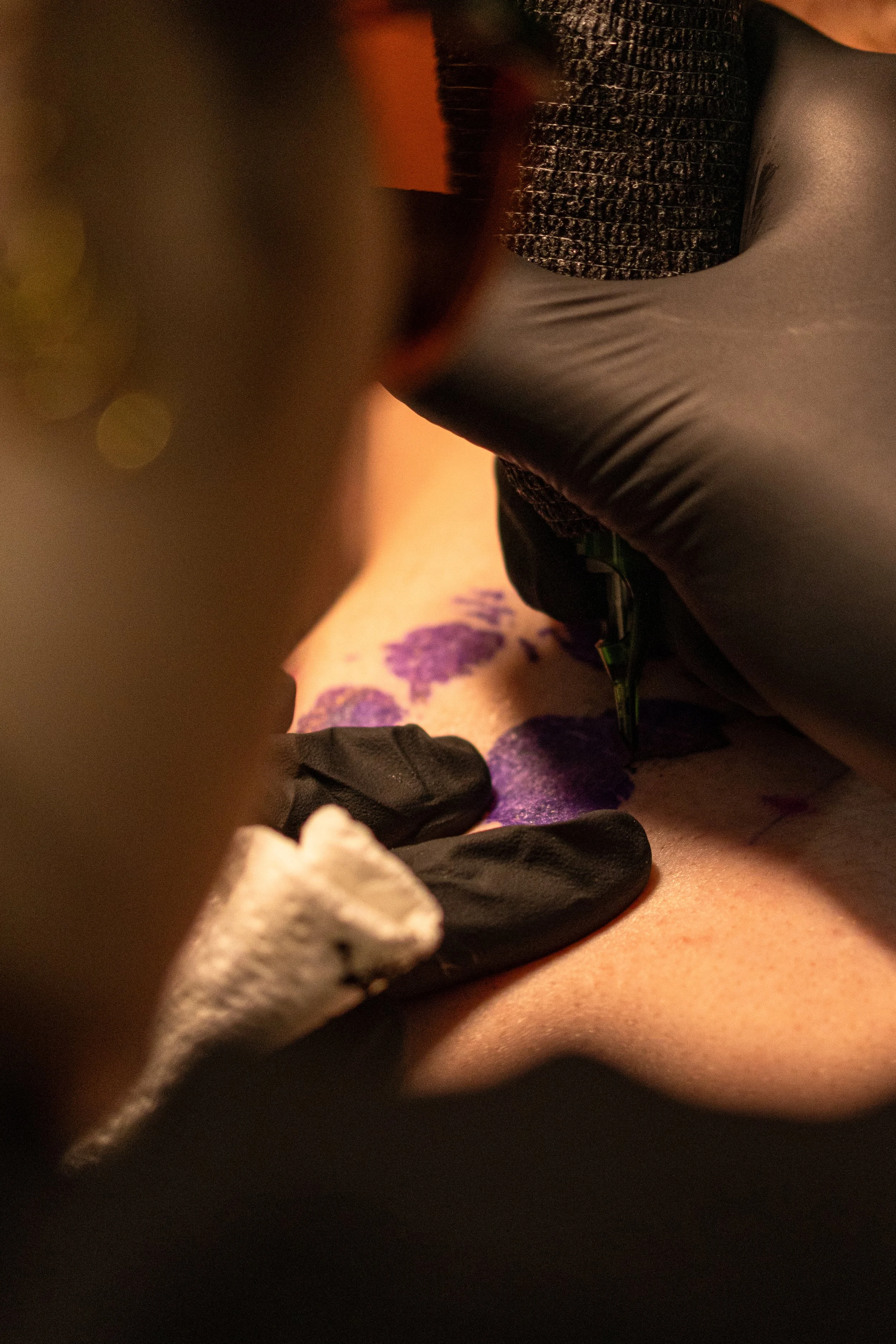 A person receiving a tattoo on their side by a tattoo artist. The artist is wearing black gloves and using a tattoo machine. The person's skin appears to have a tattoo stencil outline in purple ink.