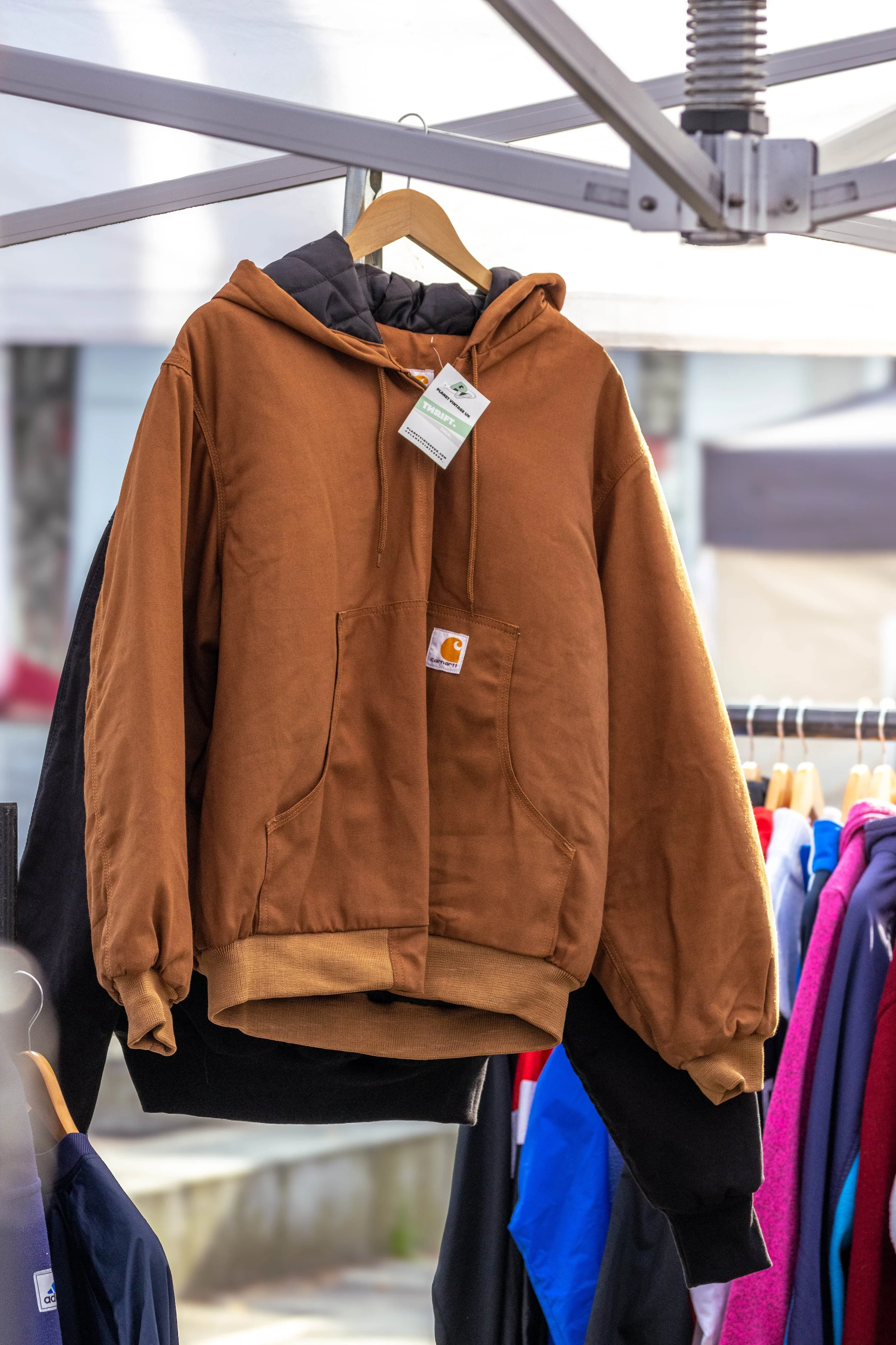 Brown Carhartt hoodie with a front pocket hanging on a wooden hanger at a clothing stall, with other colorful clothing items visible on racks nearby.