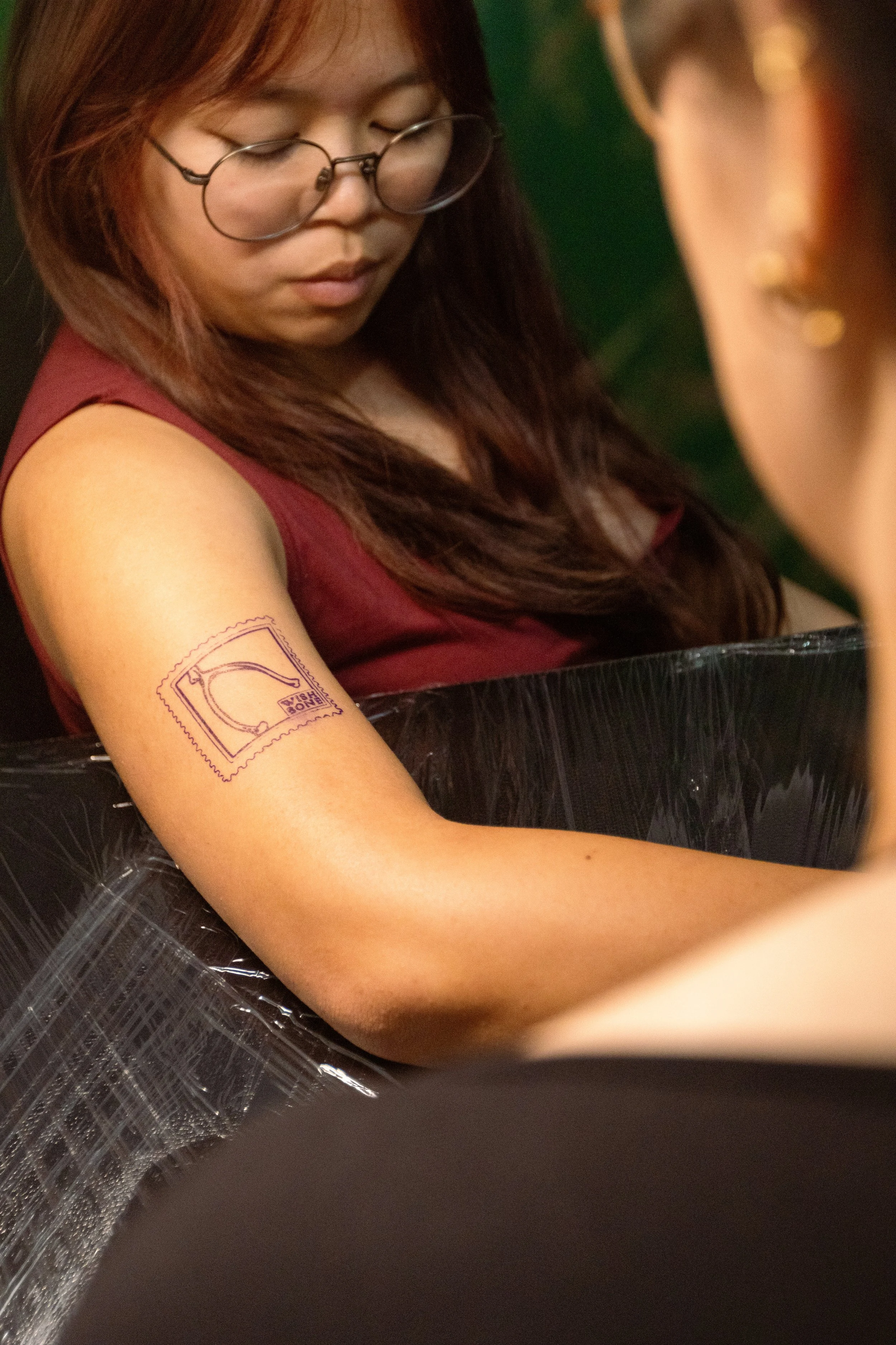 A woman with long brown hair, wearing glasses and a sleeveless top, is looking down. She has a temporary tattoo of a postage stamp on her upper arm and is sitting on black plastic with some green background behind her.