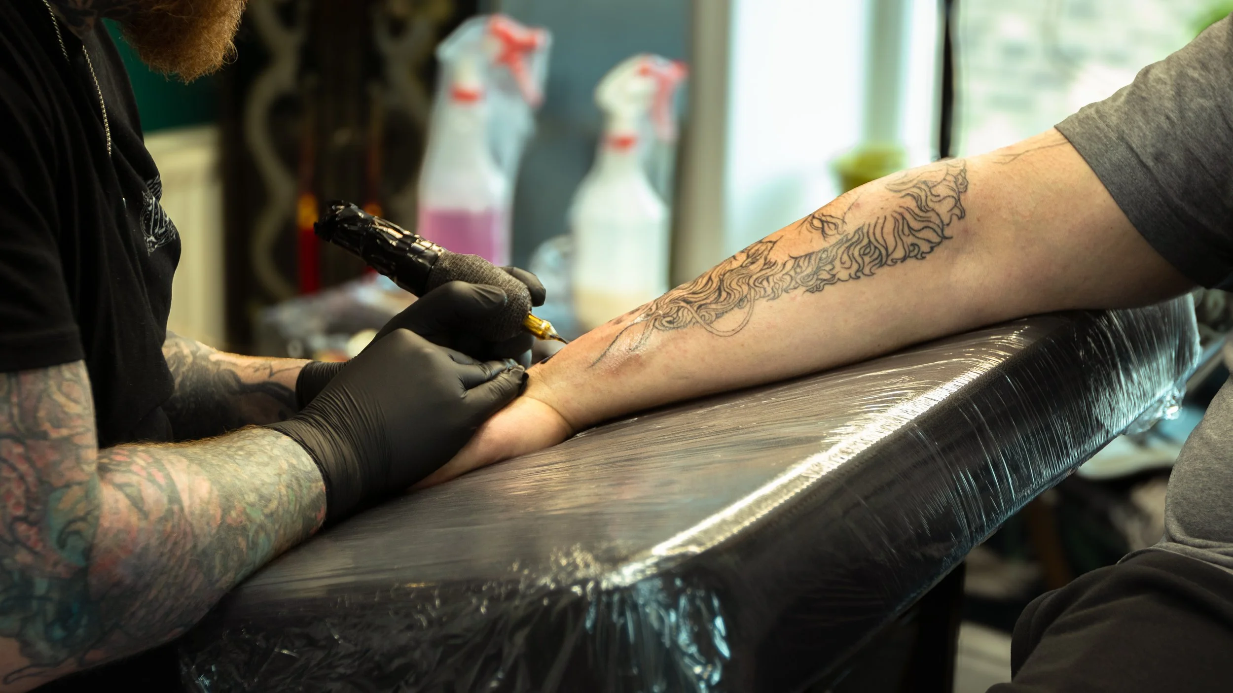 A tattoo artist is tattooing a design on a person's forearm in a tattoo studio.