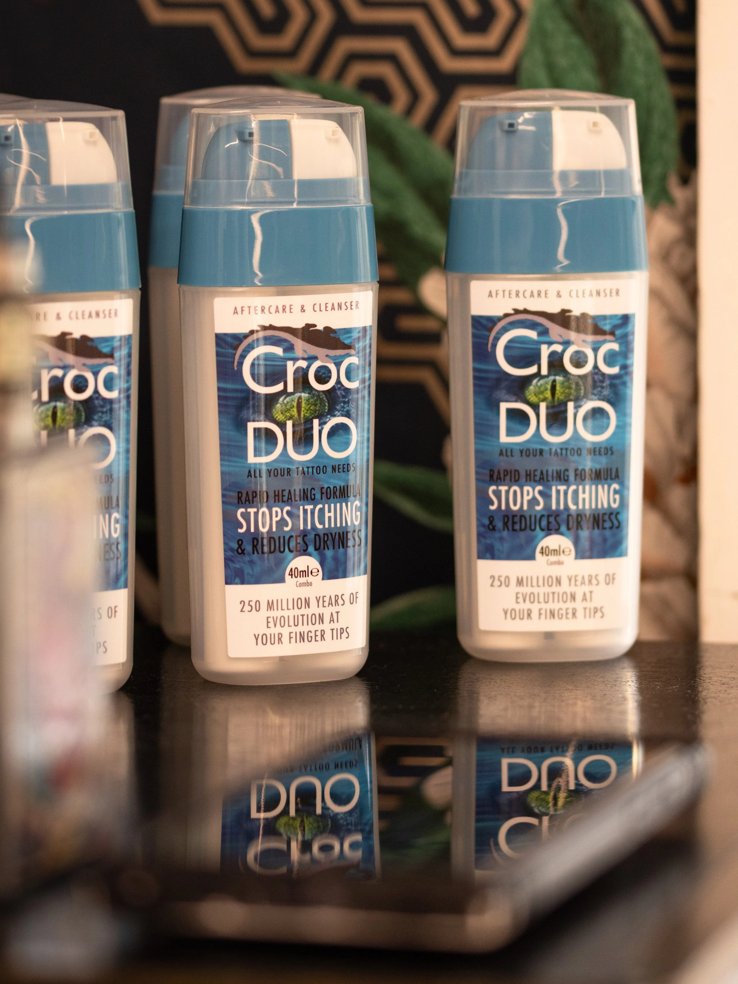 Multiple bottles of Croc Duo aftercare and cleanser standing upright on a surface, with a dark patterned background and some green leaves partially visible behind them.
