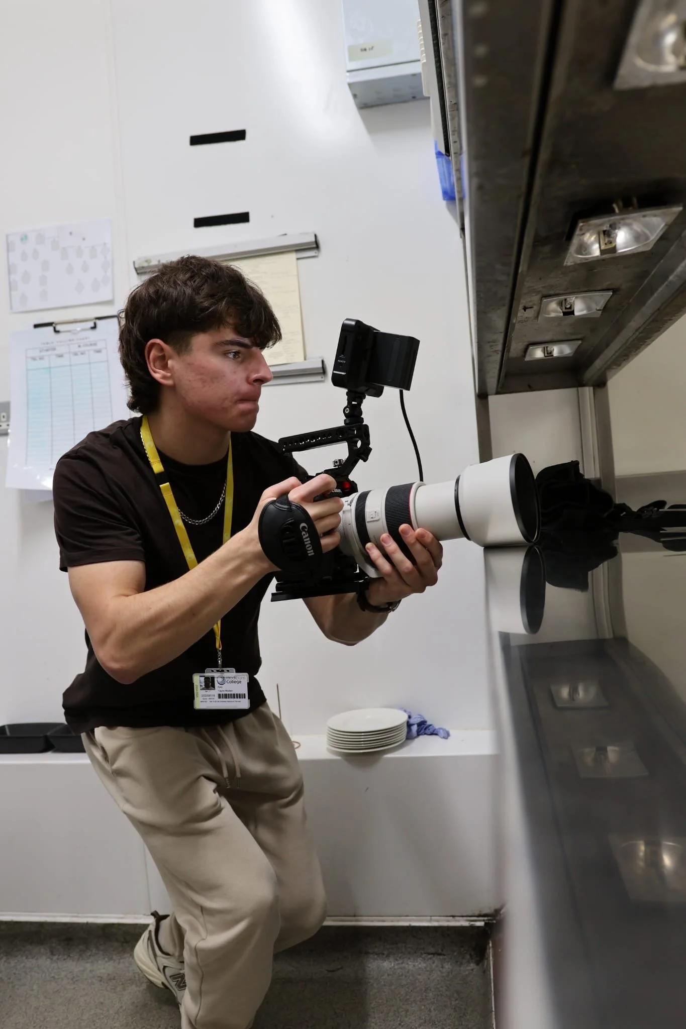 A young man holding a professional camera with a large telephoto lens while kneeling in a kitchen area, looking intently at the camera screen.