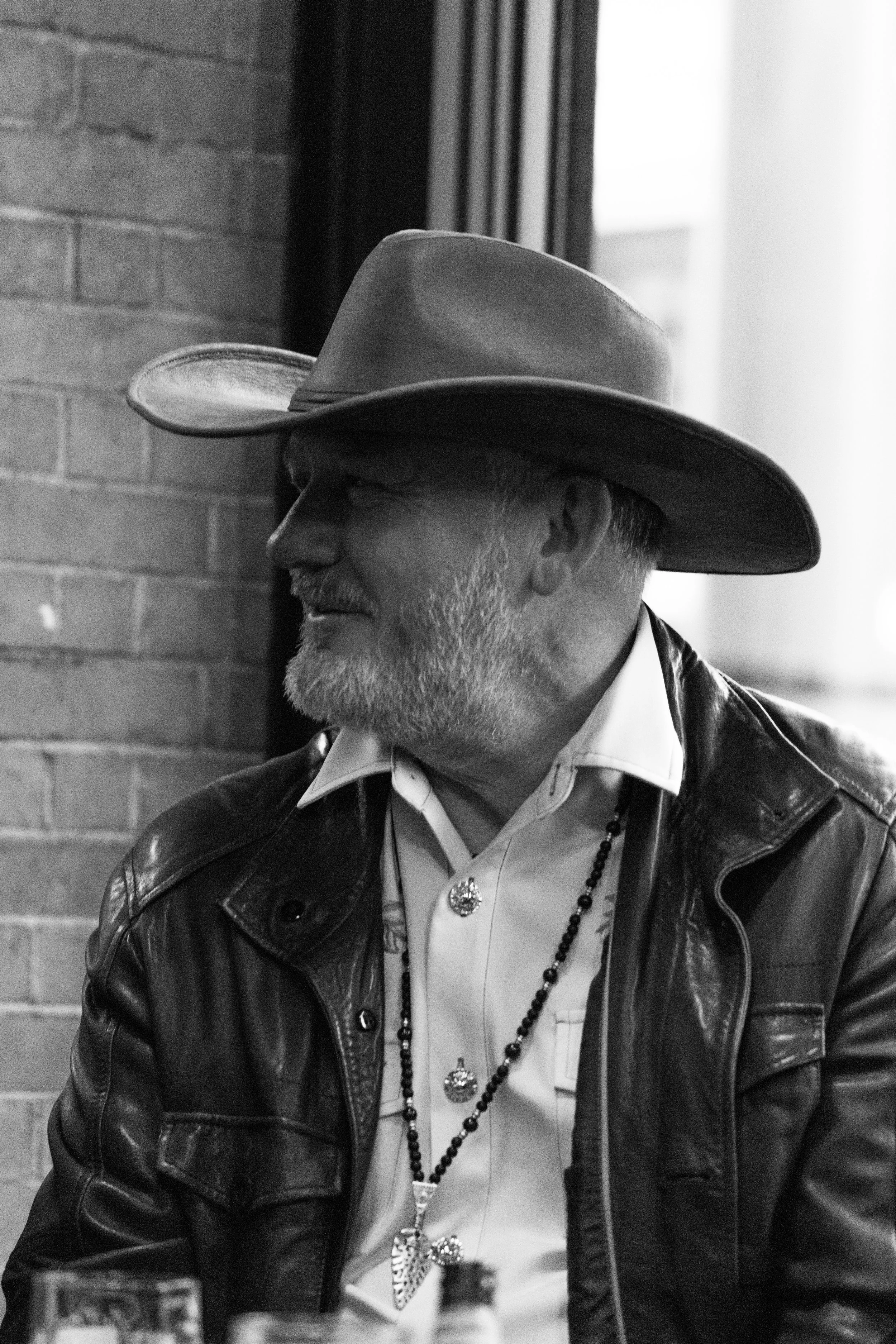 A man with a beard wearing glasses, a cowboy hat, a leather jacket, a white shirt, and several necklaces, including one with a butterfly pendant, is sitting indoors near a brick wall and a window.