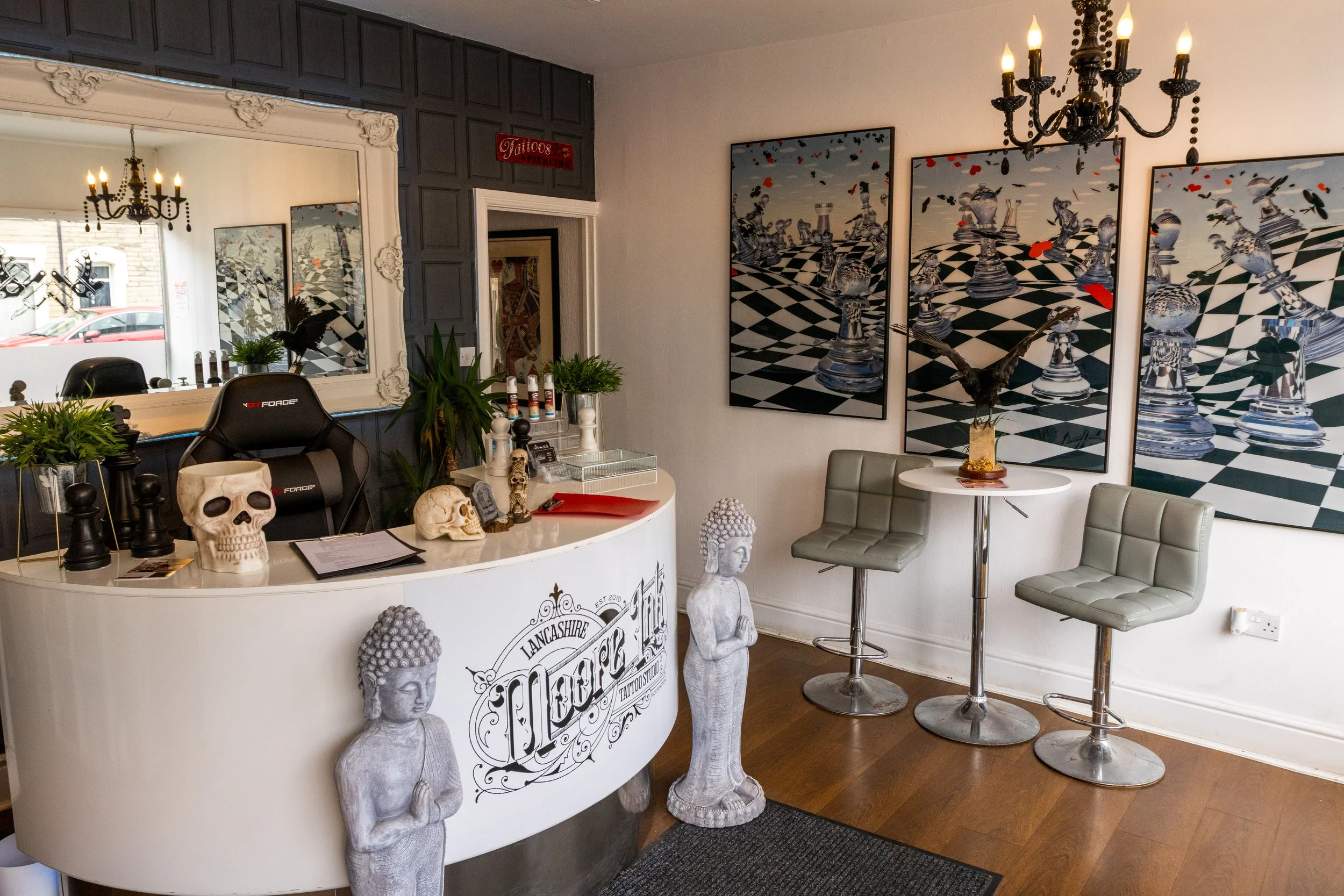 Interior of a tattoo studio reception area with a white curved desk, skull sculptures, greenery, and a large ornate mirror. There are three gray bar stools and framed chess-themed artwork on the wall. Decorative statues and a chandelier hang from the