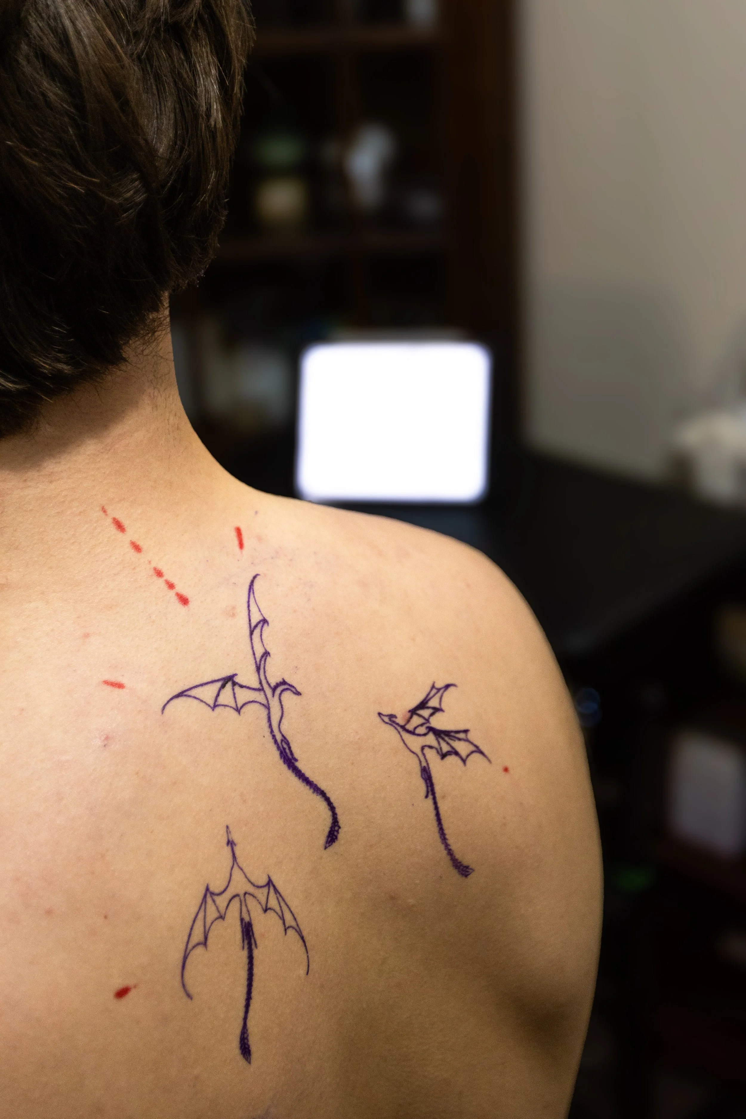 Close-up of a person's shoulder and back with colorful temporary tattoos of bats, spiders, and a spider web, with a blurred computer monitor in the background.
