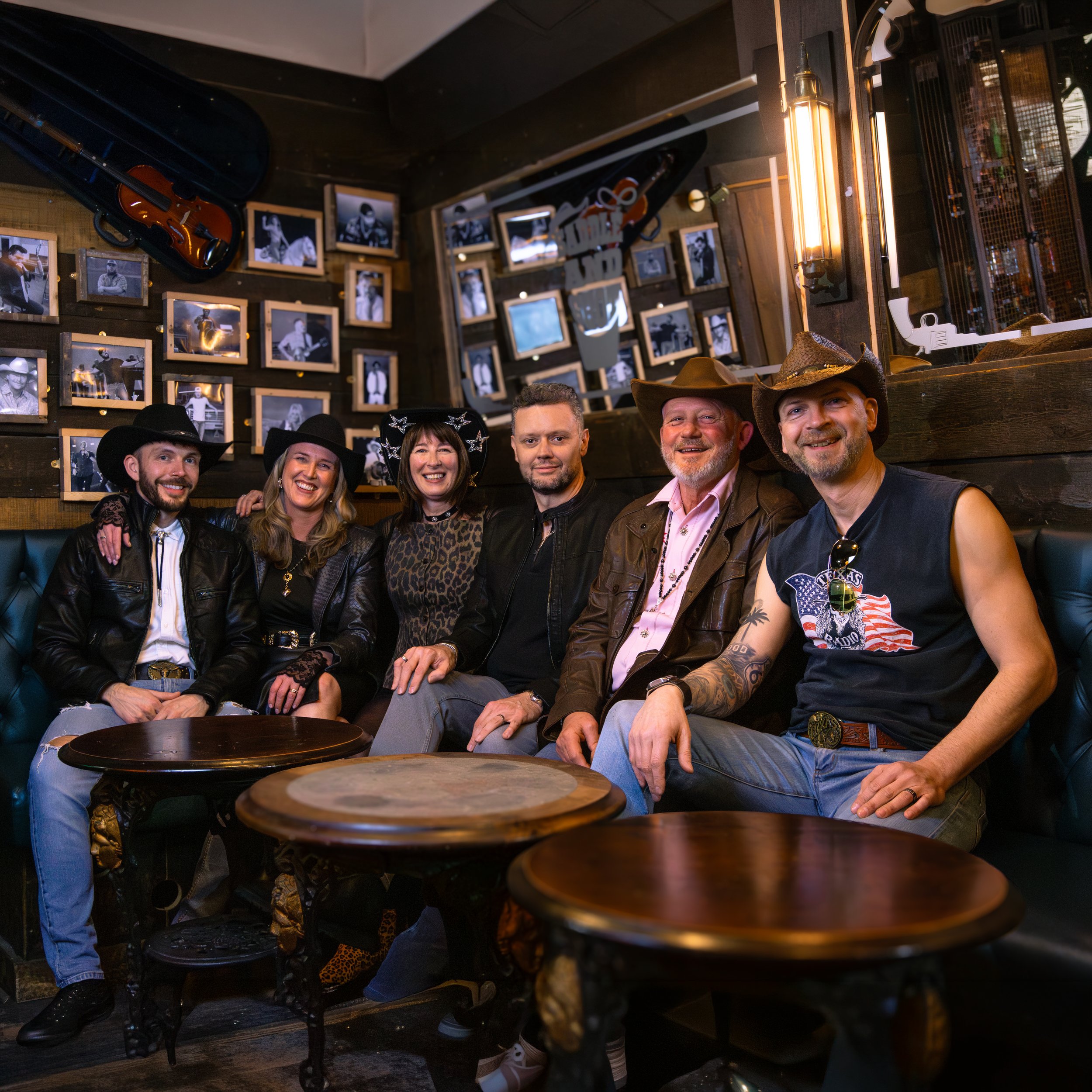 A group of six smiling people, four men and two women, dressed in cowboy attire, sitting in a dimly lit restaurant or bar with dark wooden walls decorated with black-and-white photos and displayed musical instruments, including a violin inside an ope