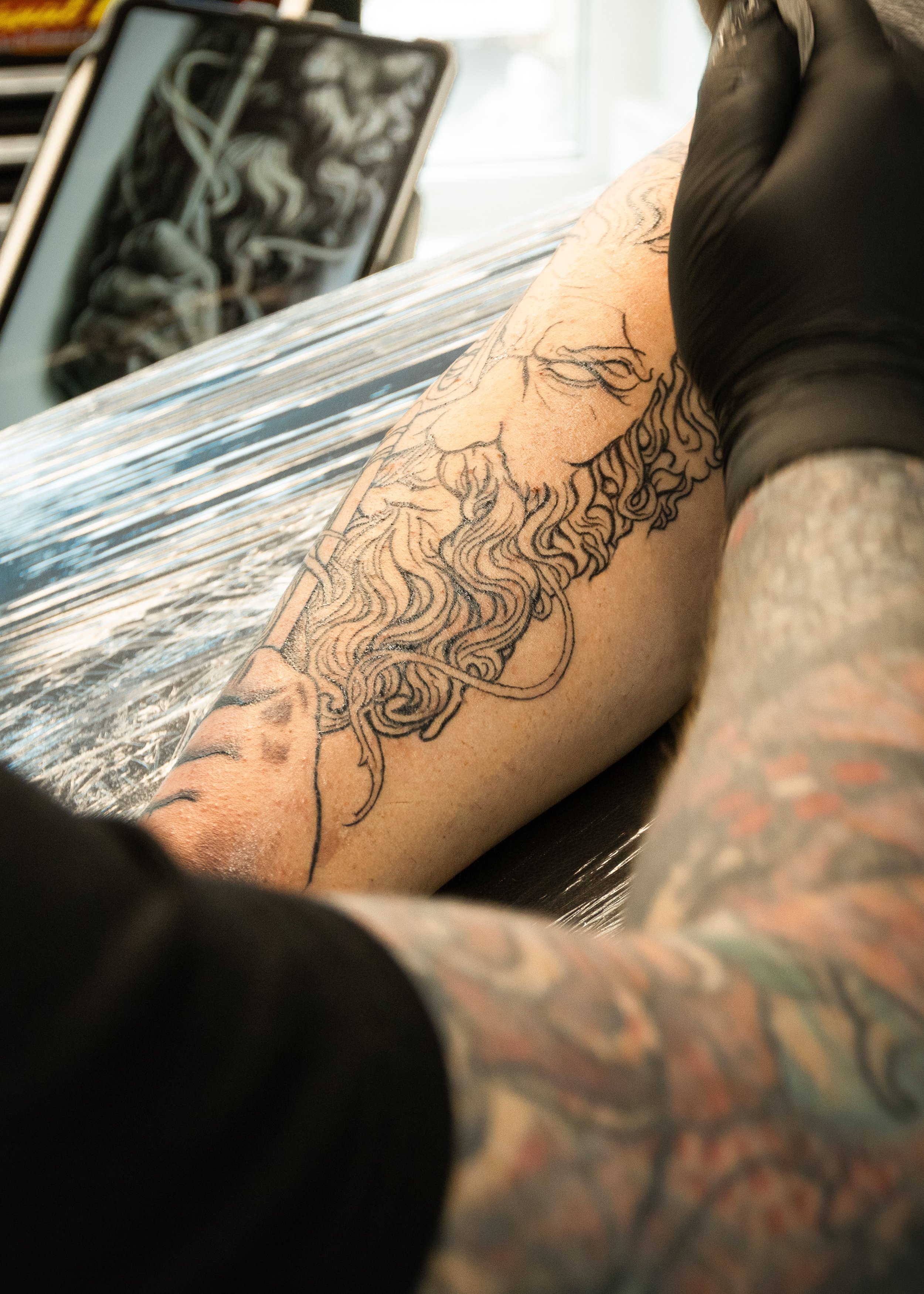 A person getting a tattoo on their arm. The tattoo is a line drawing of a bearded face with long hair.