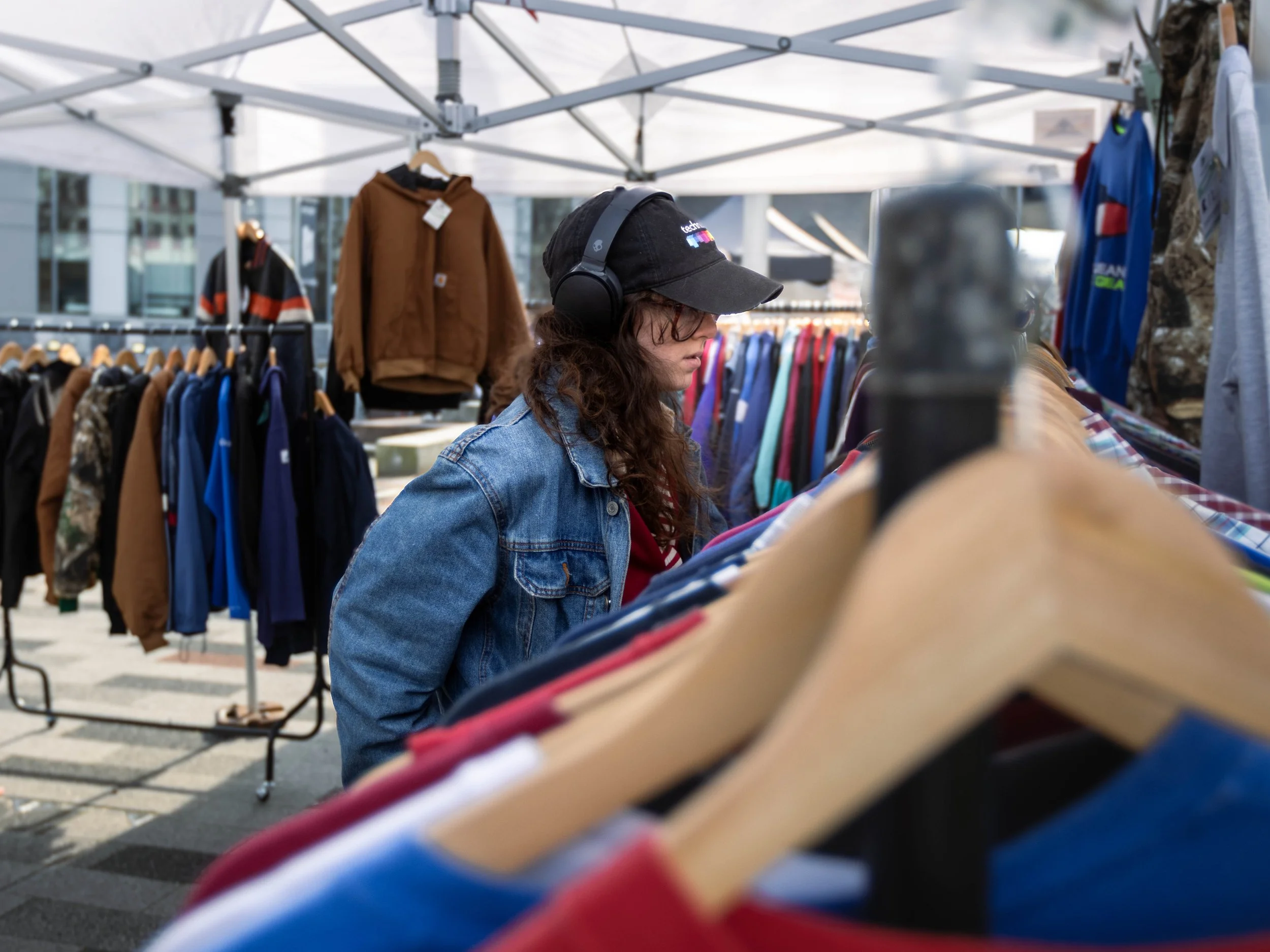 Person with long curly hair wearing a black cap and glasses shopping for clothes at an outdoor market, with racks of clothing behind them and a metal frame tent overhead.