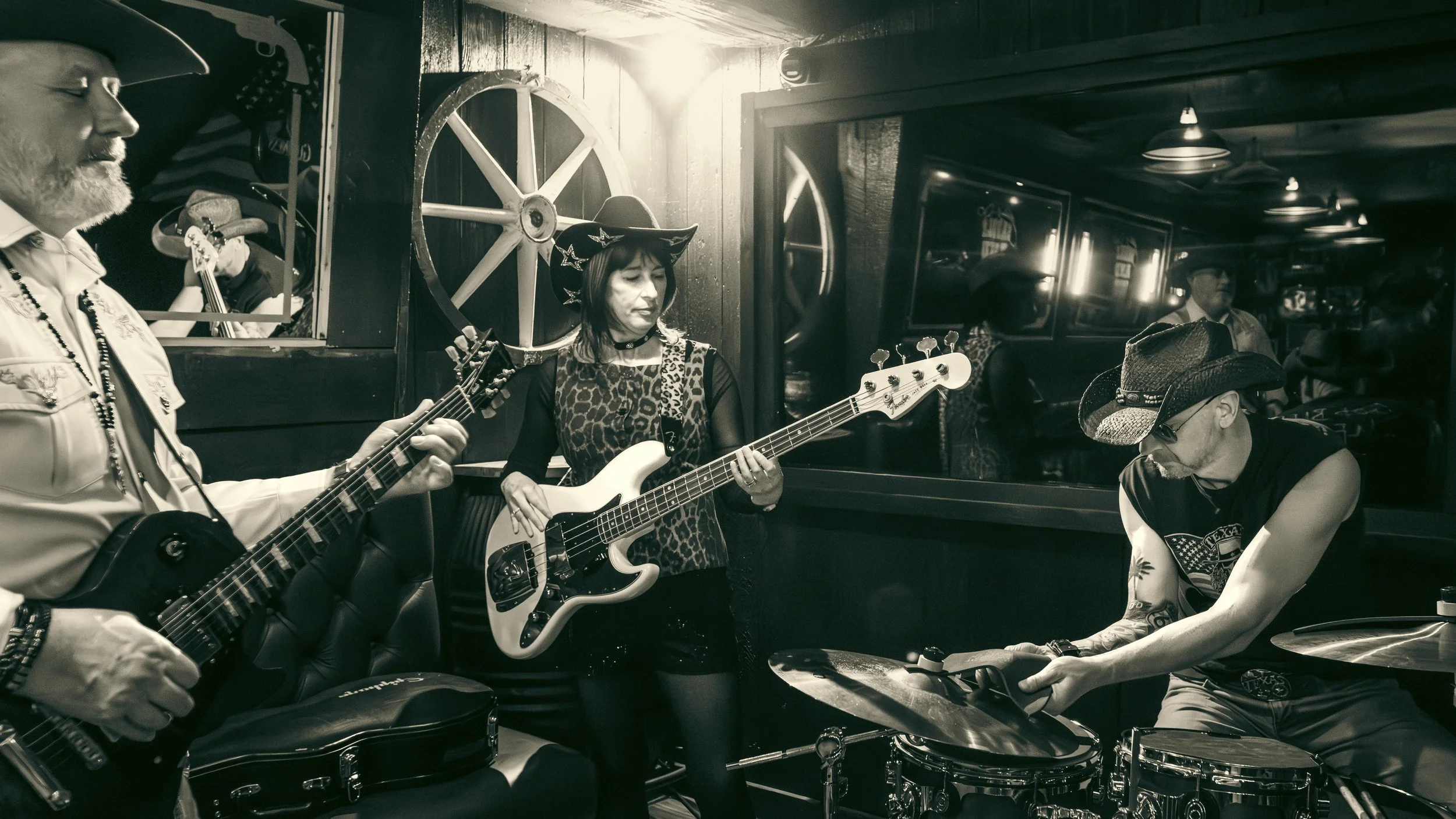 A black and white photo of a group of musicians in a dimly lit room, playing guitars and drums, with a woman in a cat hat playing a bass guitar.