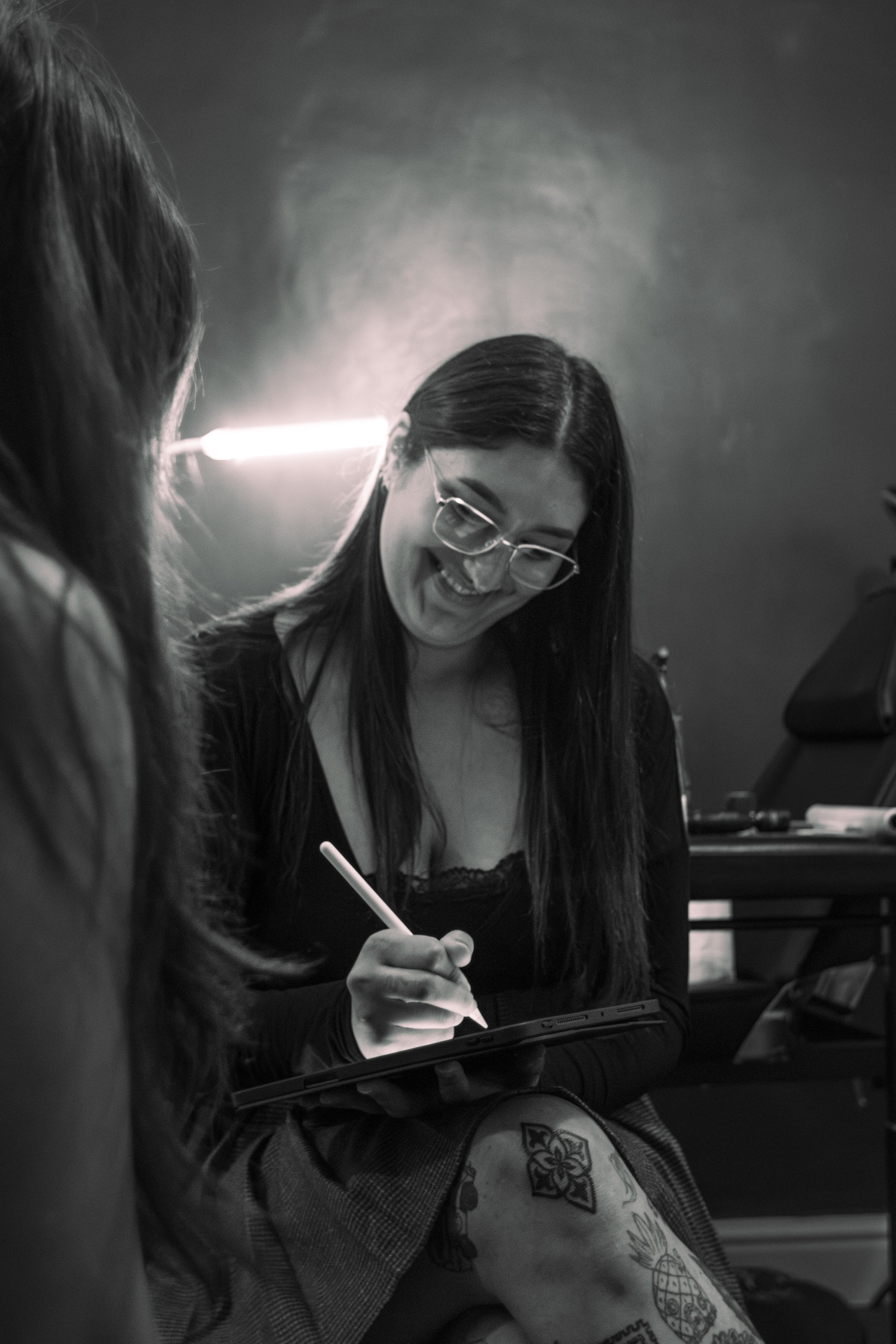 A woman with long dark hair and glasses smiling while drawing with a stylus on a digital tablet, sitting next to another person with long brown hair. The scene is black and white.