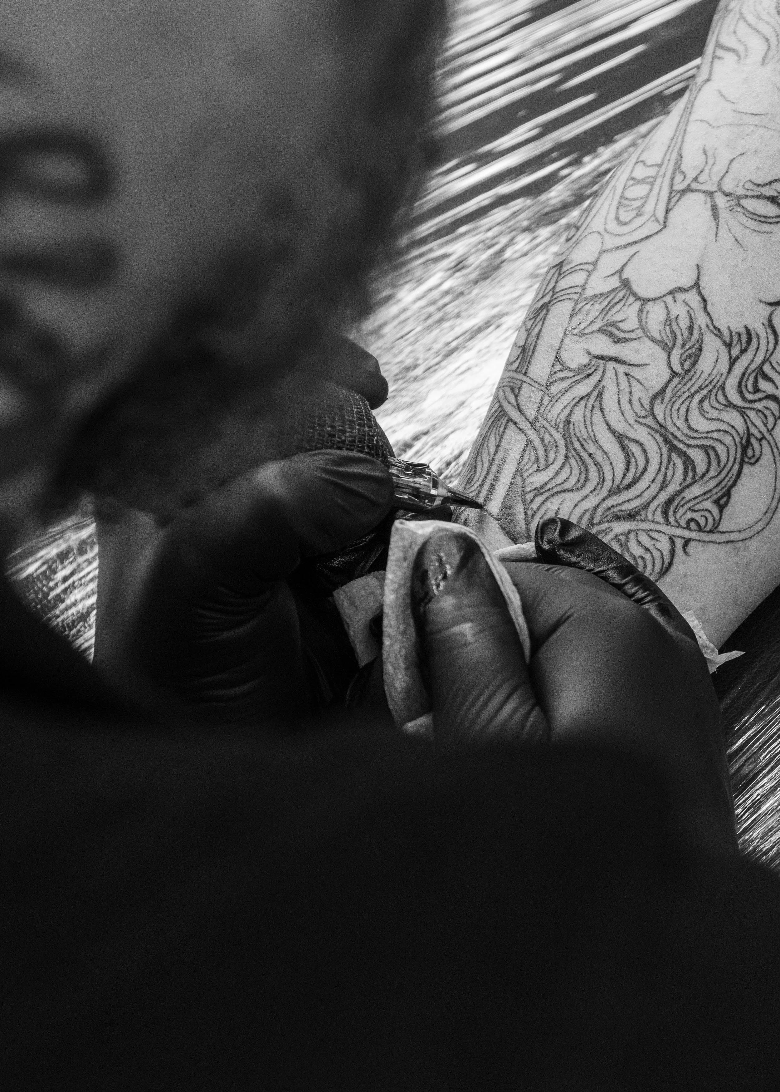 A person with gloves creates a detailed tattoo on a person's back, featuring a face with glasses and a beard with flowing hair.
