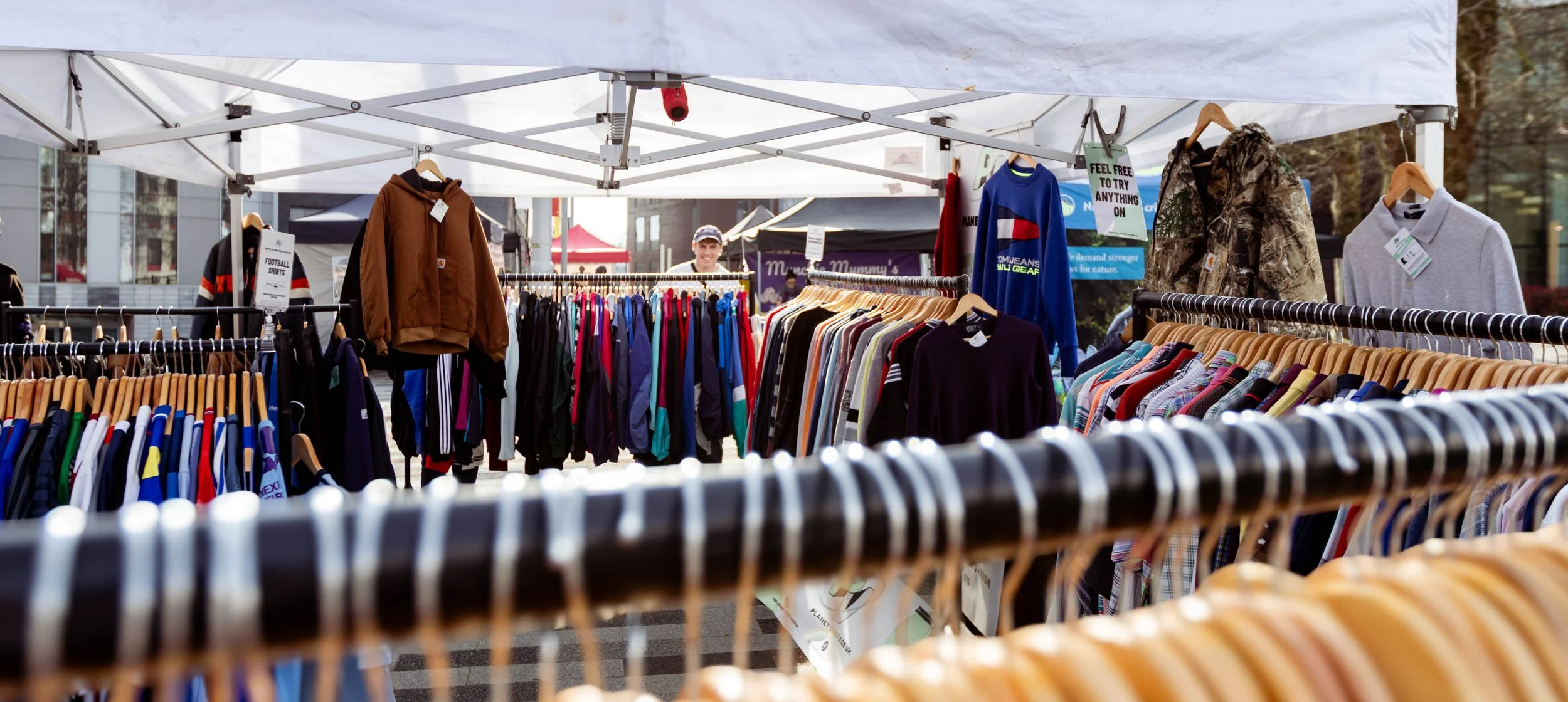 Clothing racks at an outdoor market with shirts, jackets, and hoodies on display, and a smiling man in the background.