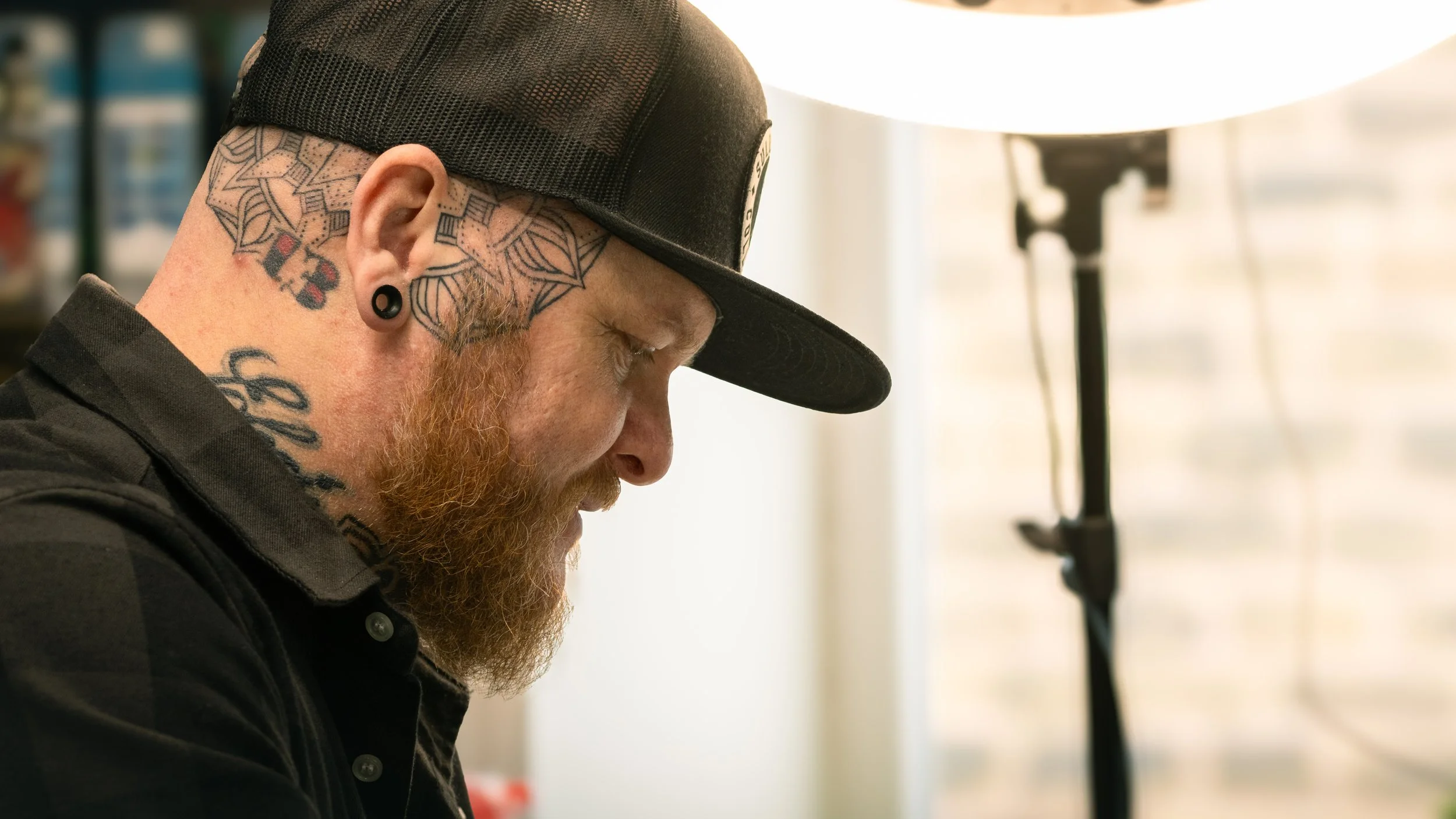 Side profile of a man with tattoos and a beard, wearing a black cap and black shirt, standing near studio lighting in an indoor setting.