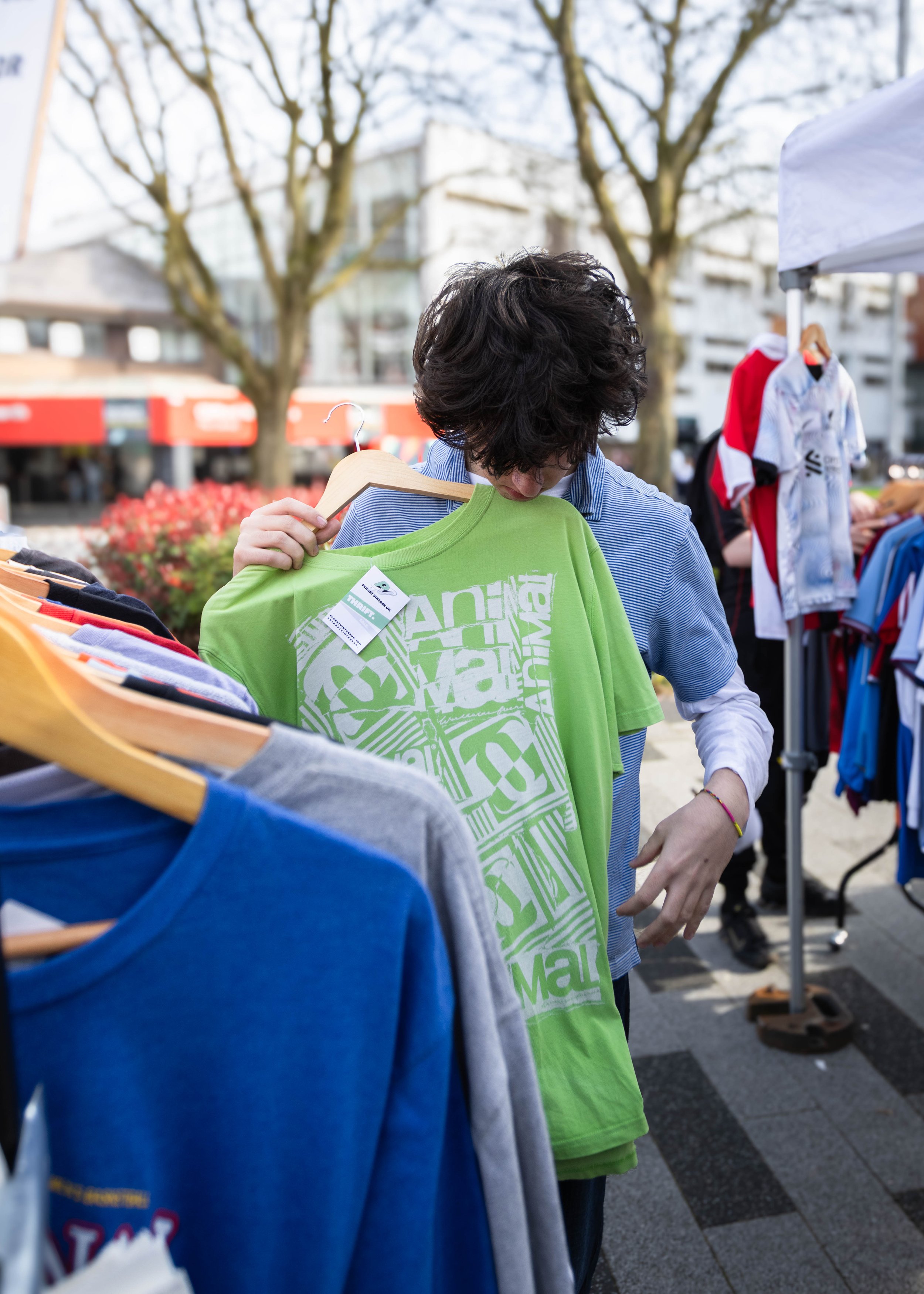 A person with curly dark hair wearing a blue shirt is shopping at an outdoor market, holding a green T-shirt with white graphic design on a hanger. Other clothing items are visible on racks nearby, and trees and buildings are in the background.