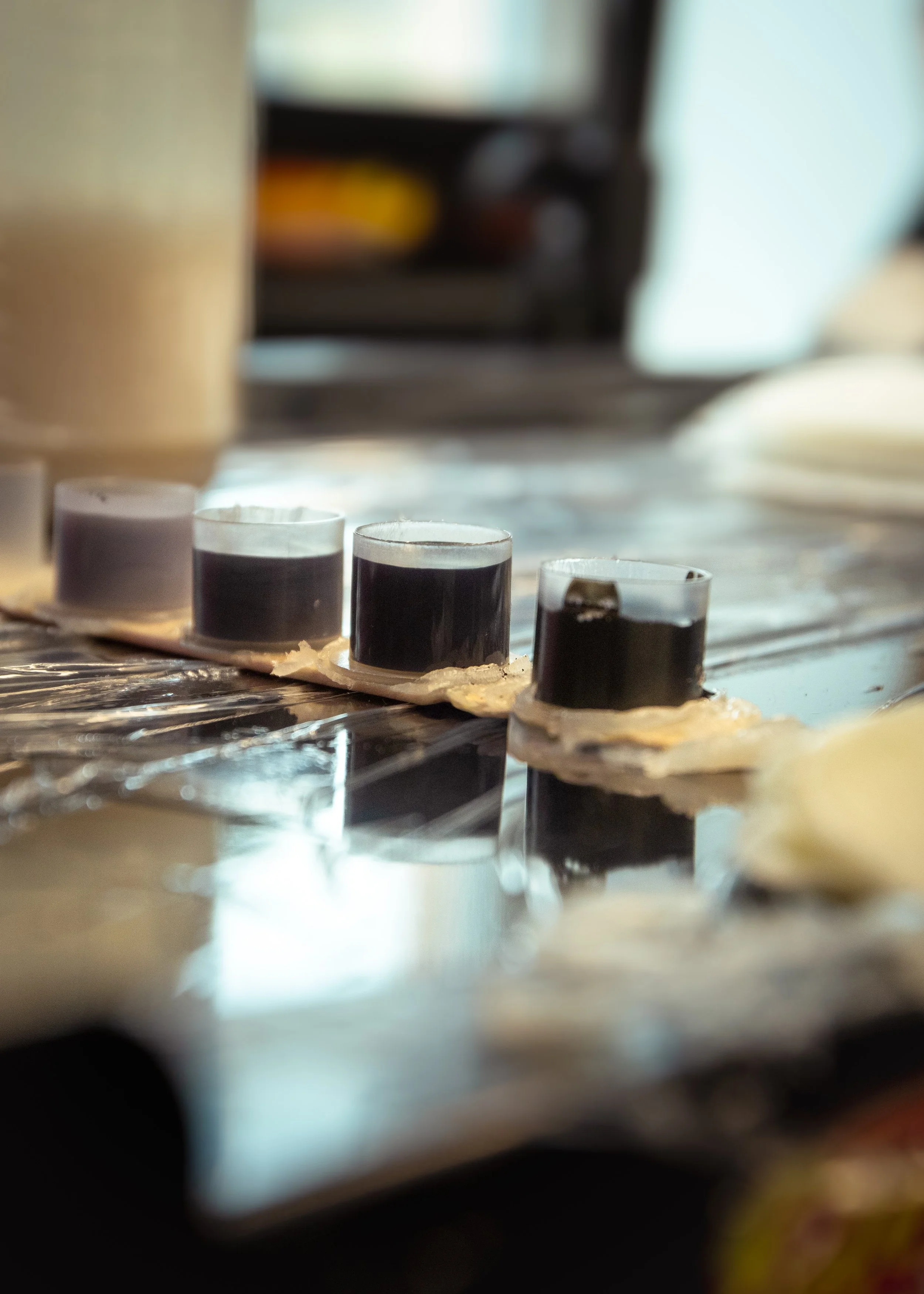 A close-up of four small shot glasses filled with dark liquid, placed on a tray covered with foil, with a blurred kitchen background.