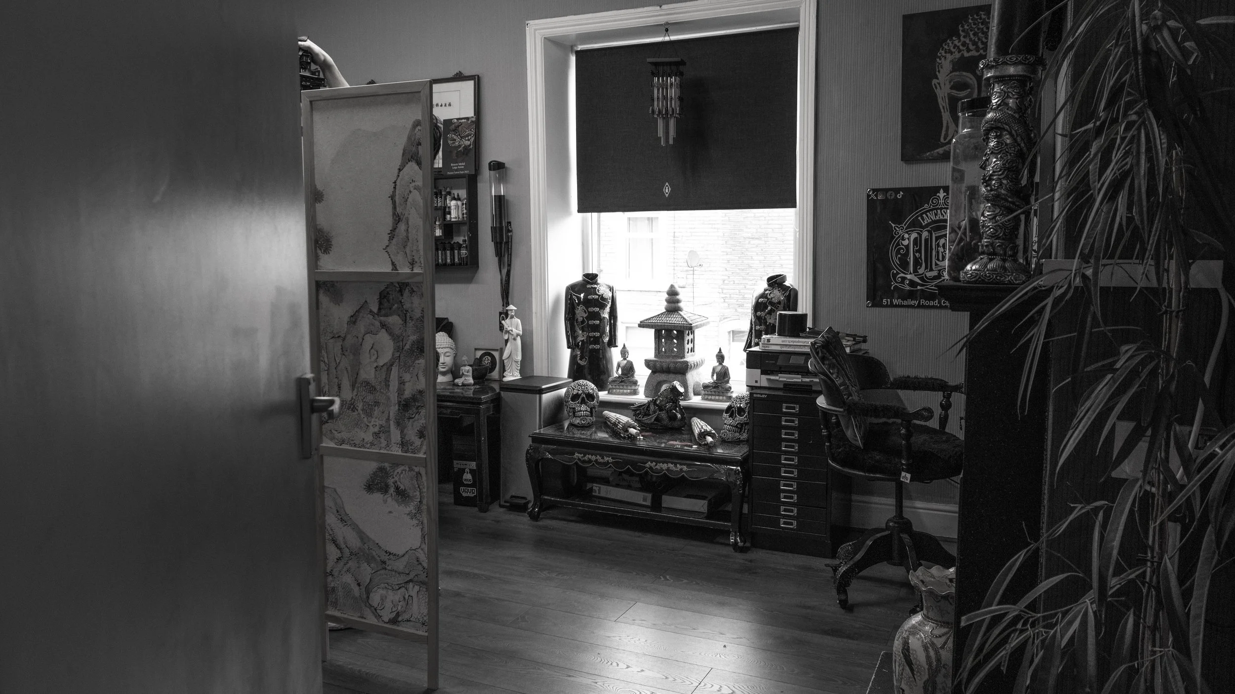 A room with various Asian and spiritual decor, including Buddha statues, skull ornaments, and a carved wooden table positioned by a window with a dark blind, and walls decorated with pictures and artwork.