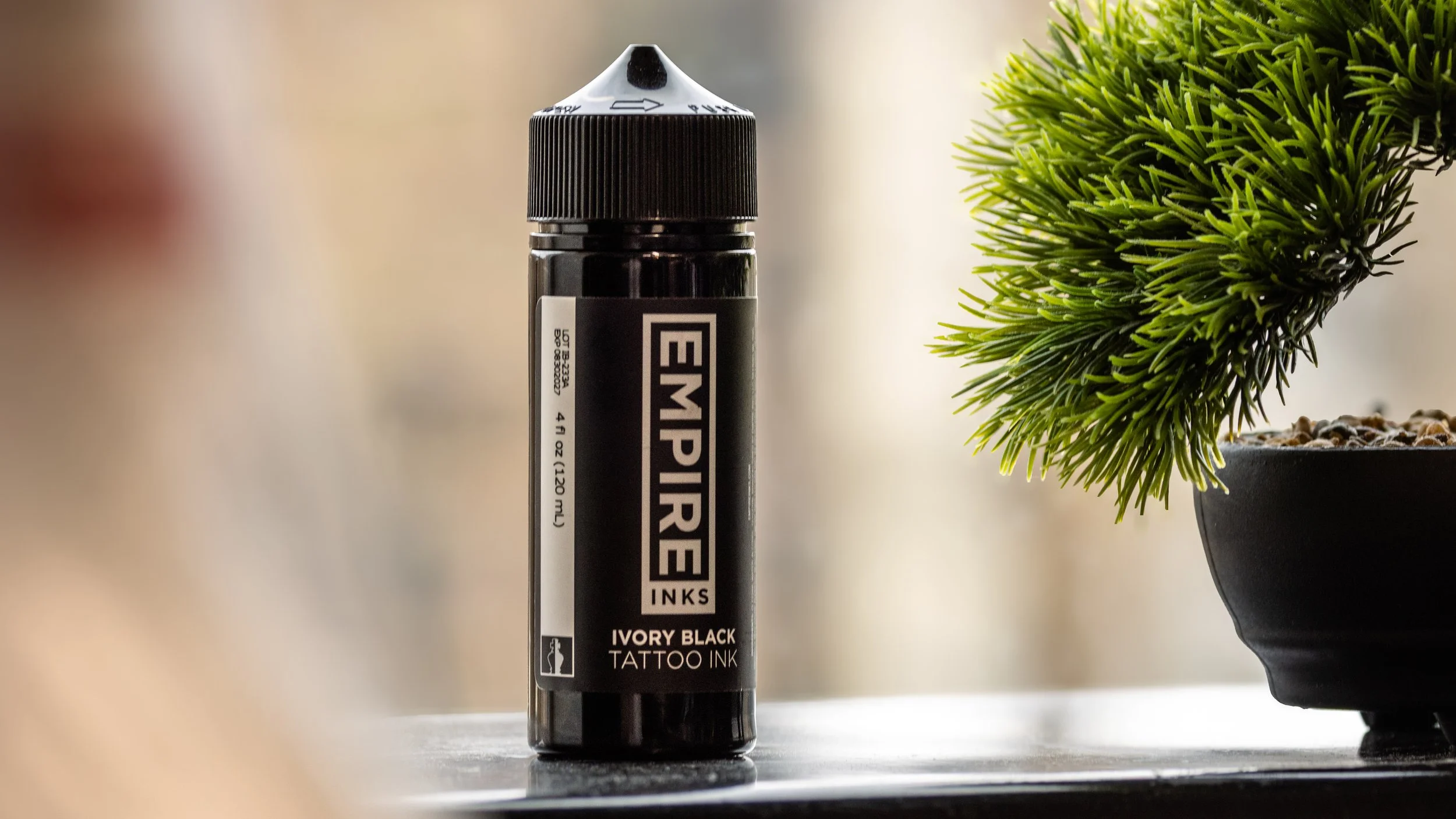 A black marker labeled "EMPIRE INKS Ivory Black Tattoo Ink" standing upright on a flat surface next to a potted green plant with needle-like leaves.