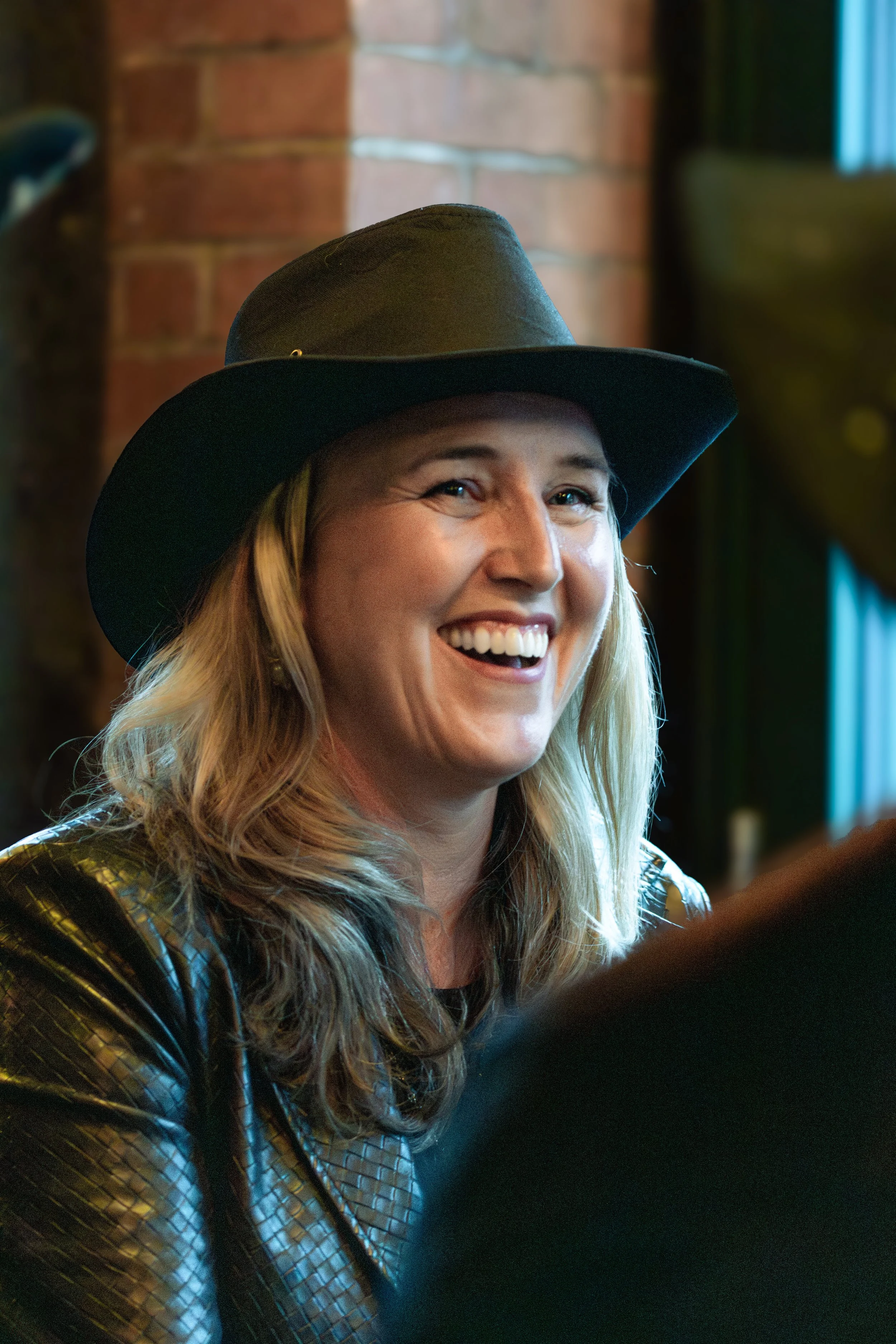 A woman with blonde hair wearing a black wide-brimmed hat, smiling, in front of a brick wall.